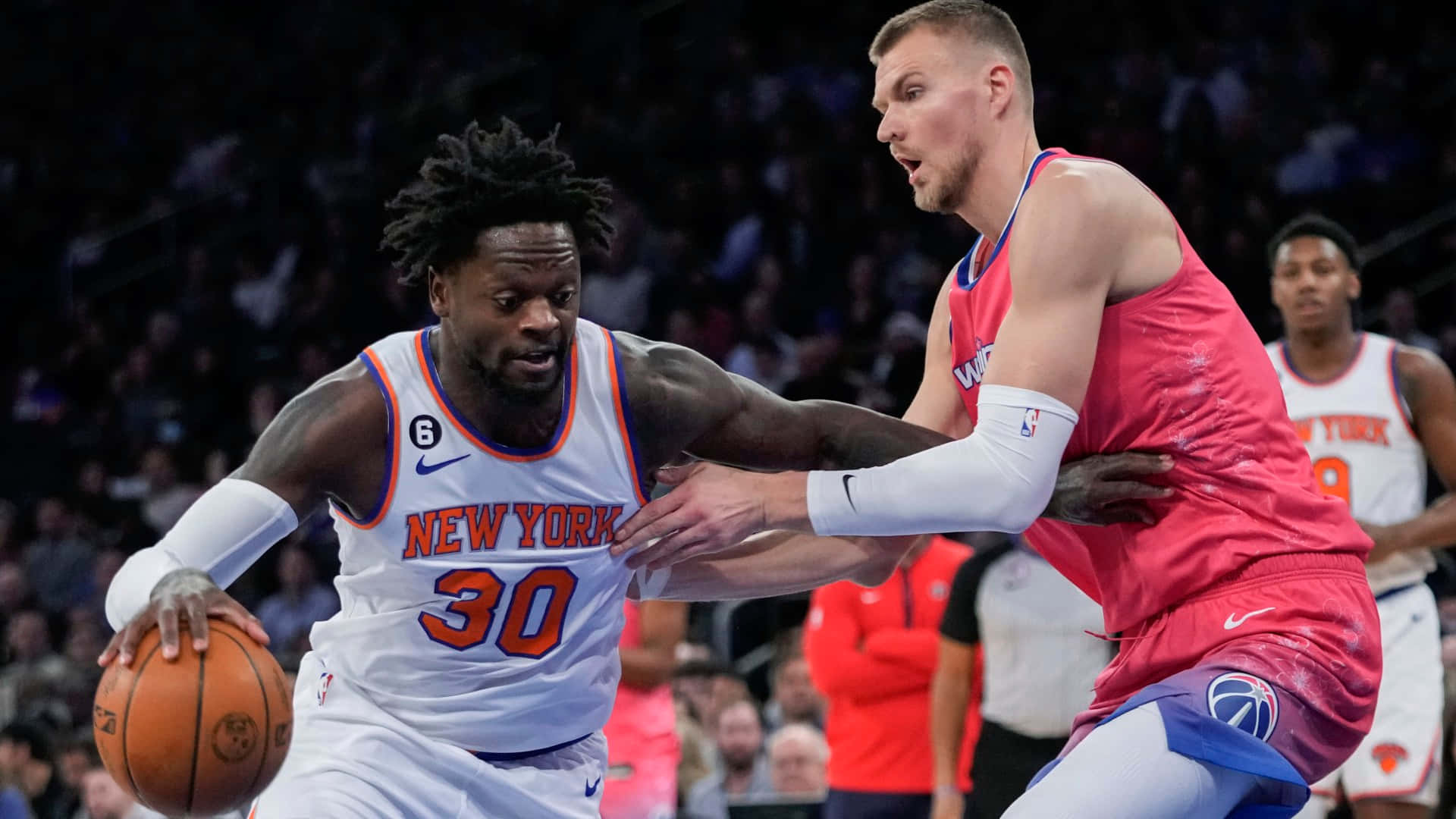 Patrick Ewing 桌布 壁紙 — 一名身穿白色紐約球衣的籃球運動員在激烈的比賽中運球，同時被一名身穿紅色球衣的高大對手緊密防守。
