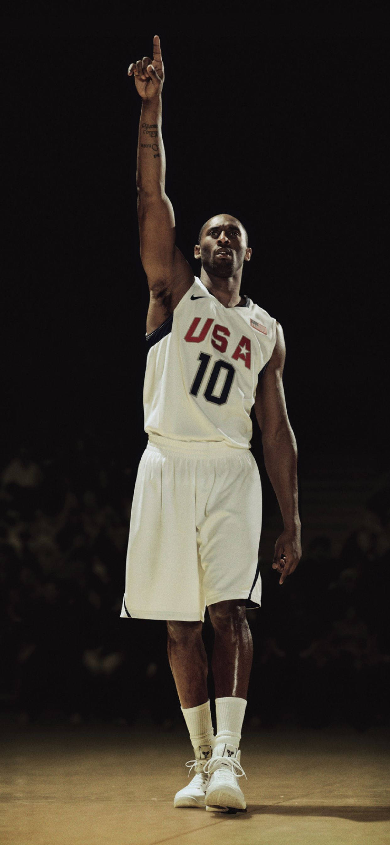 Kobe Bryant iPhone 桌布 壁紙 — 一名身穿白色美國隊10號球衣的籃球運動員在燈光昏暗的球場上舉起右手向上指。