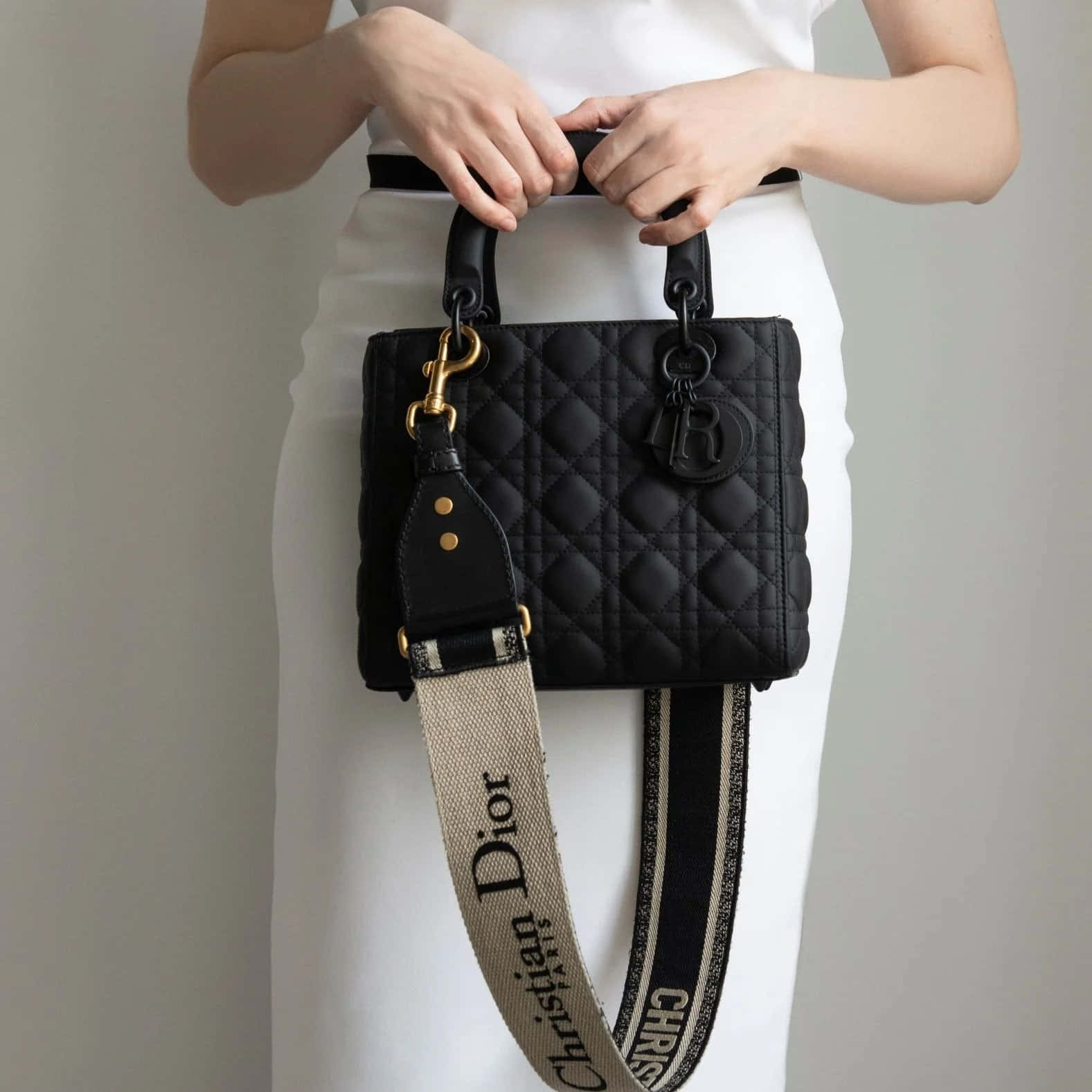 Dior 動態 桌布 壁紙 — 一名身穿白色衣服的人，在純色背景前拿著一個黑色絎縫Dior手袋，配有金色金屬配件和品牌肩帶。