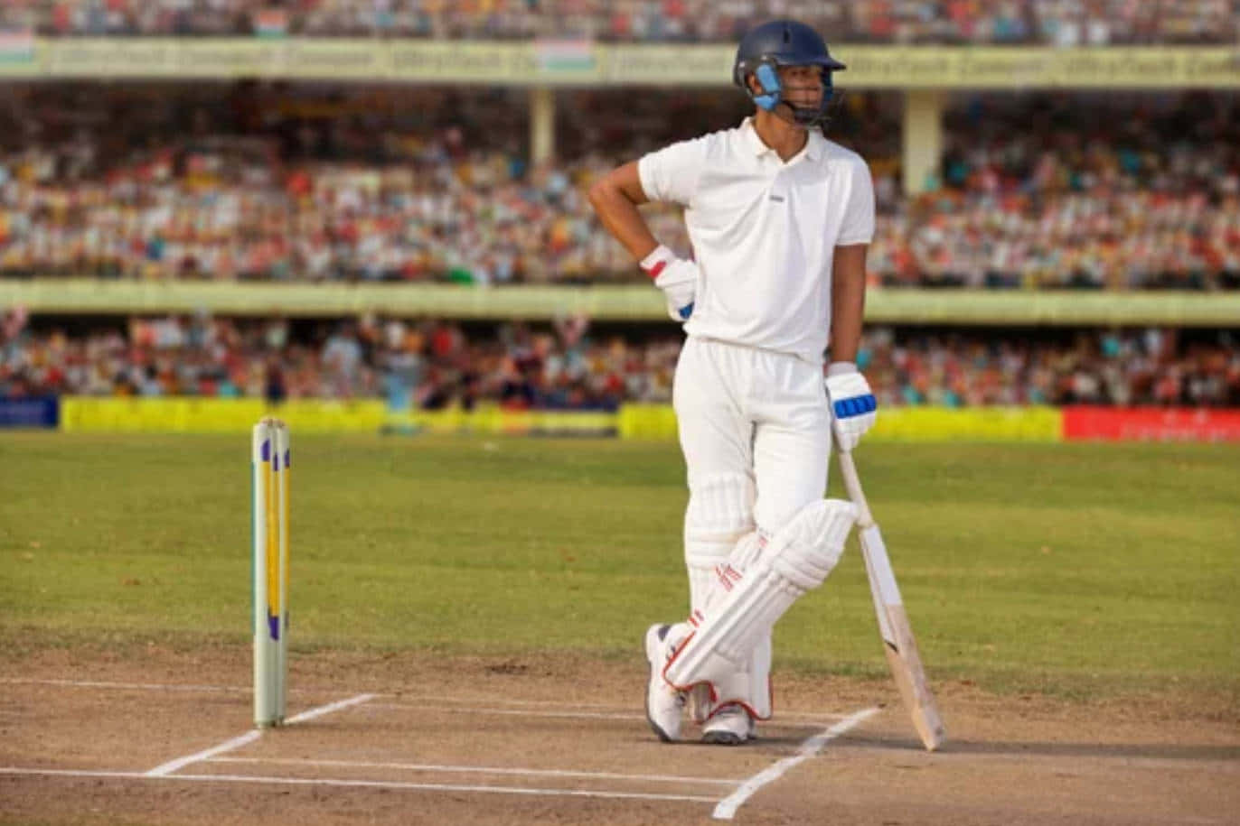Cricket Ground 桌布 壁紙 — 一名身穿白色裝備的板球擊球手站在陽光普照的綠色球場的擊球線上，手持球棒，面對座無虛席的體育場。