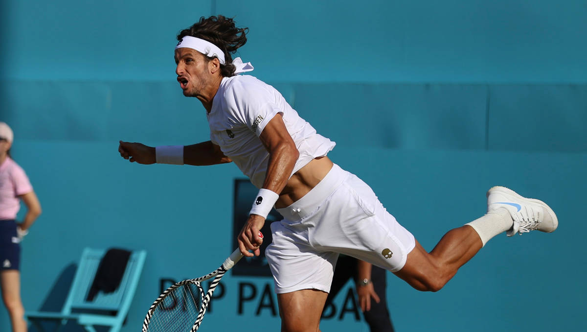 Tennis 桌布 壁紙 — 一名身穿白色裝備的男性網球運動員在揮拍時動態地向前衝刺，背景是明亮的藍色球場牆壁。