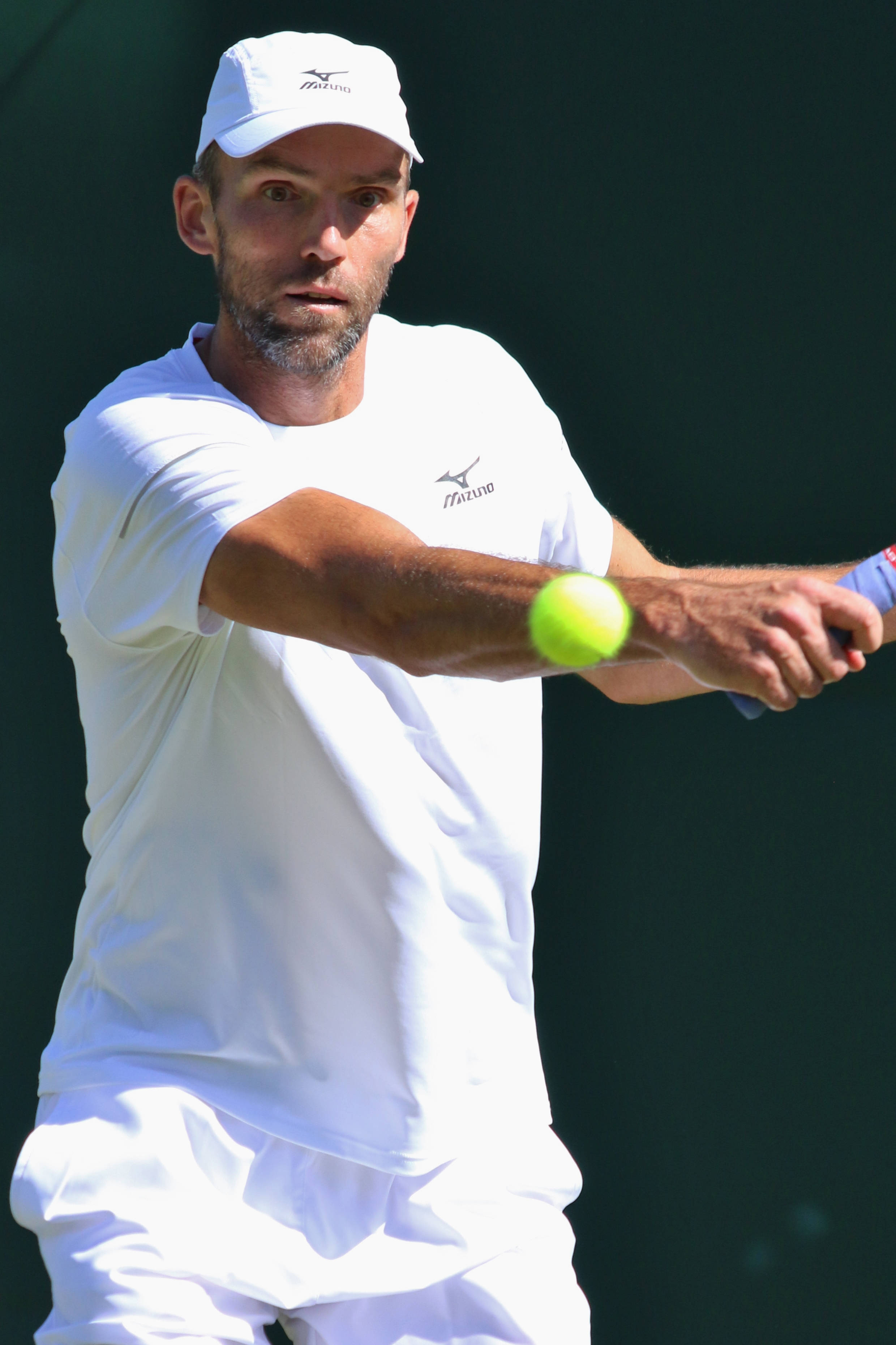 Tennis 桌布 壁紙 — 一名身穿白色裝備的男性網球選手在深綠色背景下揮拍擊打一個亮黃色的球。