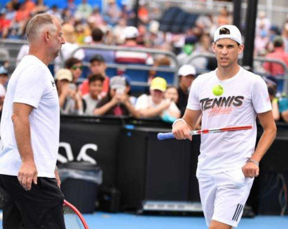 Tennis 桌布 壁紙 — 一名身穿白色裝備的網球選手拿著球拍和球，在藍色球場上與教練交談，背景有觀眾。