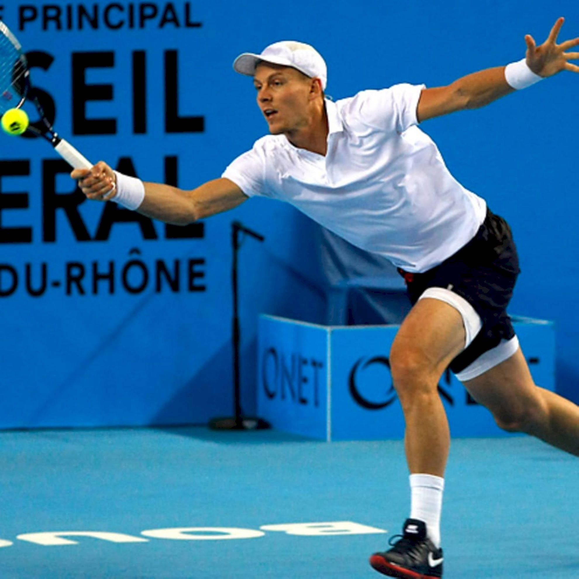 Tennis 桌布 壁紙 — 一名身穿白色襯衫和帽子的男性網球選手在藍色球場上向前撲身，用球拍擊打一個黃色球。