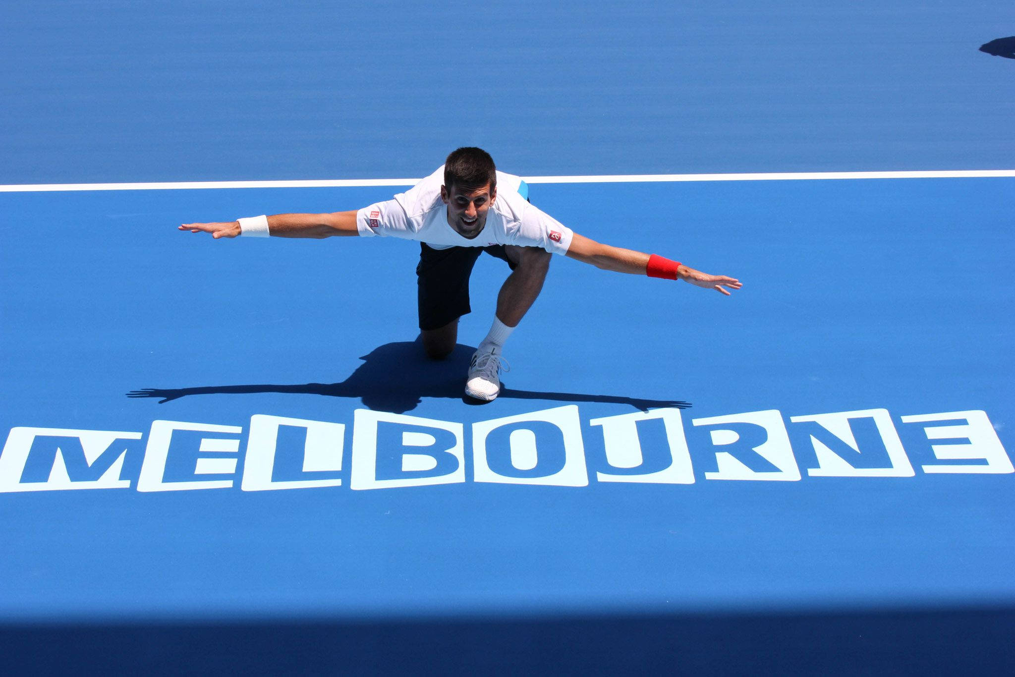 Australian Open 桌布 壁紙 — 一名身穿白色襯衫和黑色短褲的網球運動員在明亮的藍色墨爾本球場上張開雙臂慶祝。
