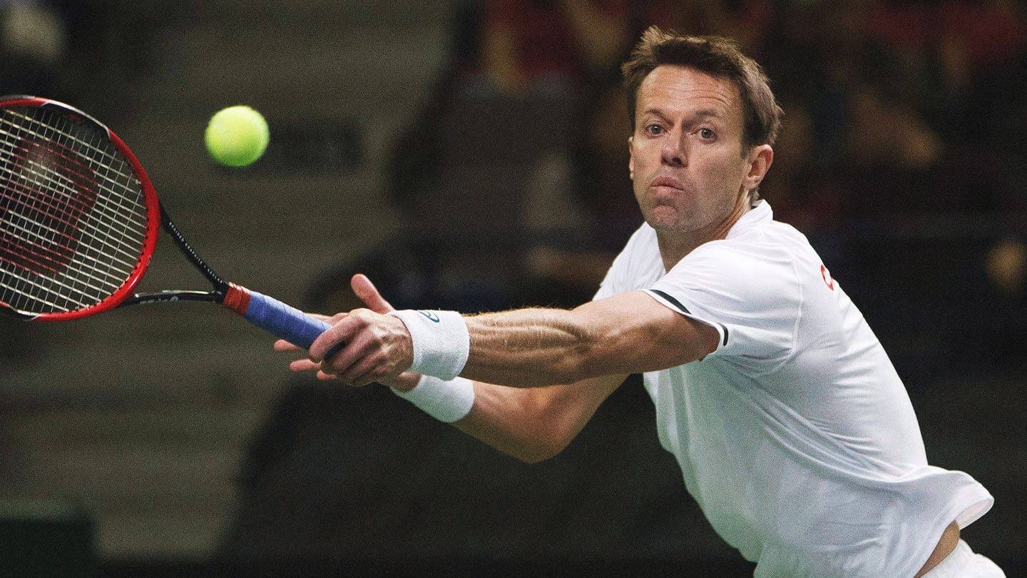 Tennis 桌布 壁紙 — 一名身穿白色襯衫的男性網球選手在室內比賽中，專注地揮動紅色球拍擊打明亮的黃色球。