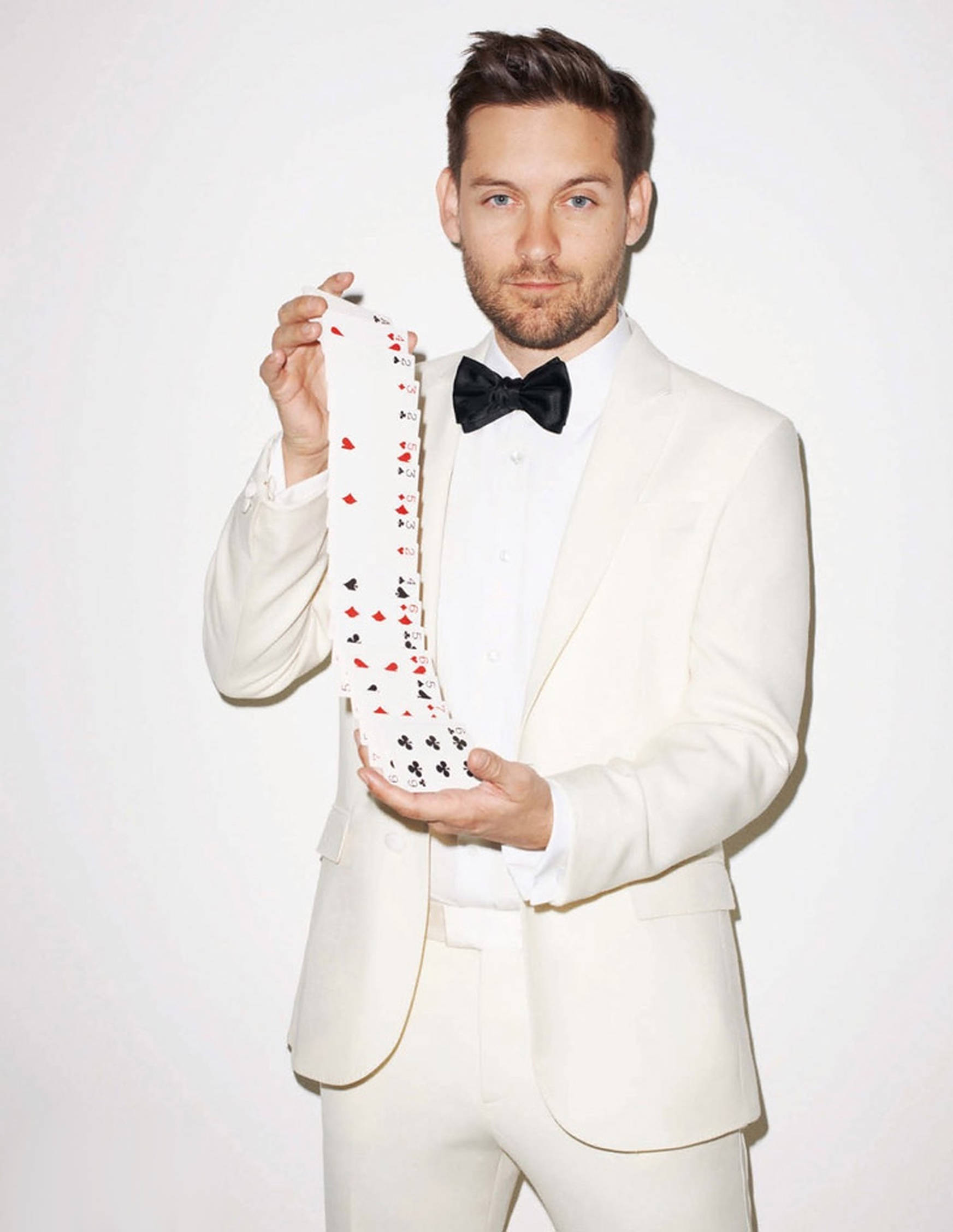 Tobey Maguire 桌布 壁紙 — 一名身穿白色西裝、戴黑色蝴蝶結領結的男子，在純白色背景前拿著一條印有撲克牌圖案的長領帶。