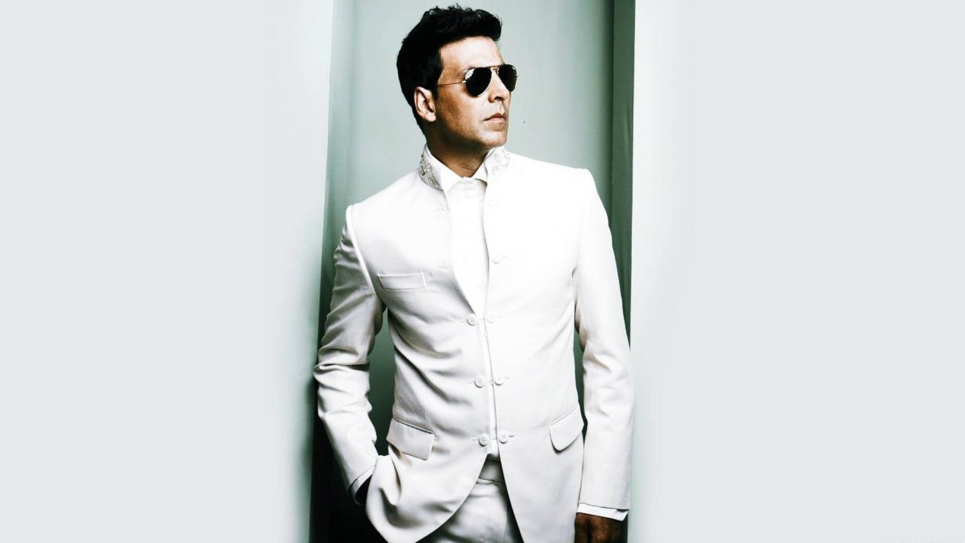 Akshay Kumar 桌布 壁紙 — 一名身穿白色西裝和太陽眼鏡的男子，自信地站在簡約淺灰色牆壁前。