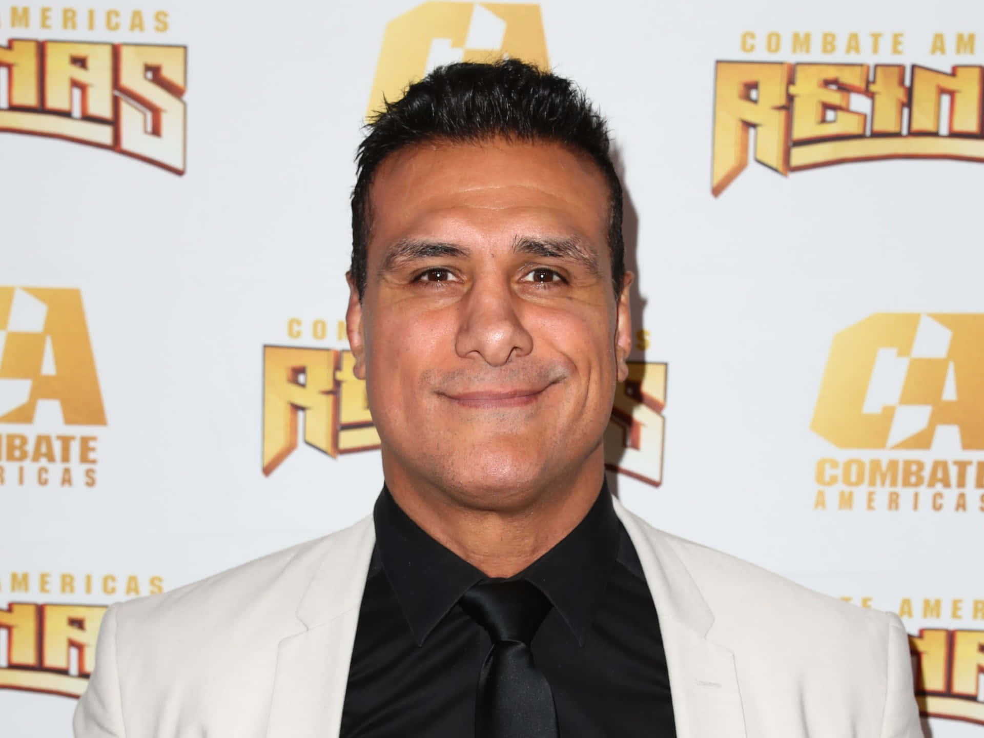 WWE 桌布 壁紙 — 一名身穿白色西裝和黑色領帶的男子在品牌活動背景前對著鏡頭微笑。