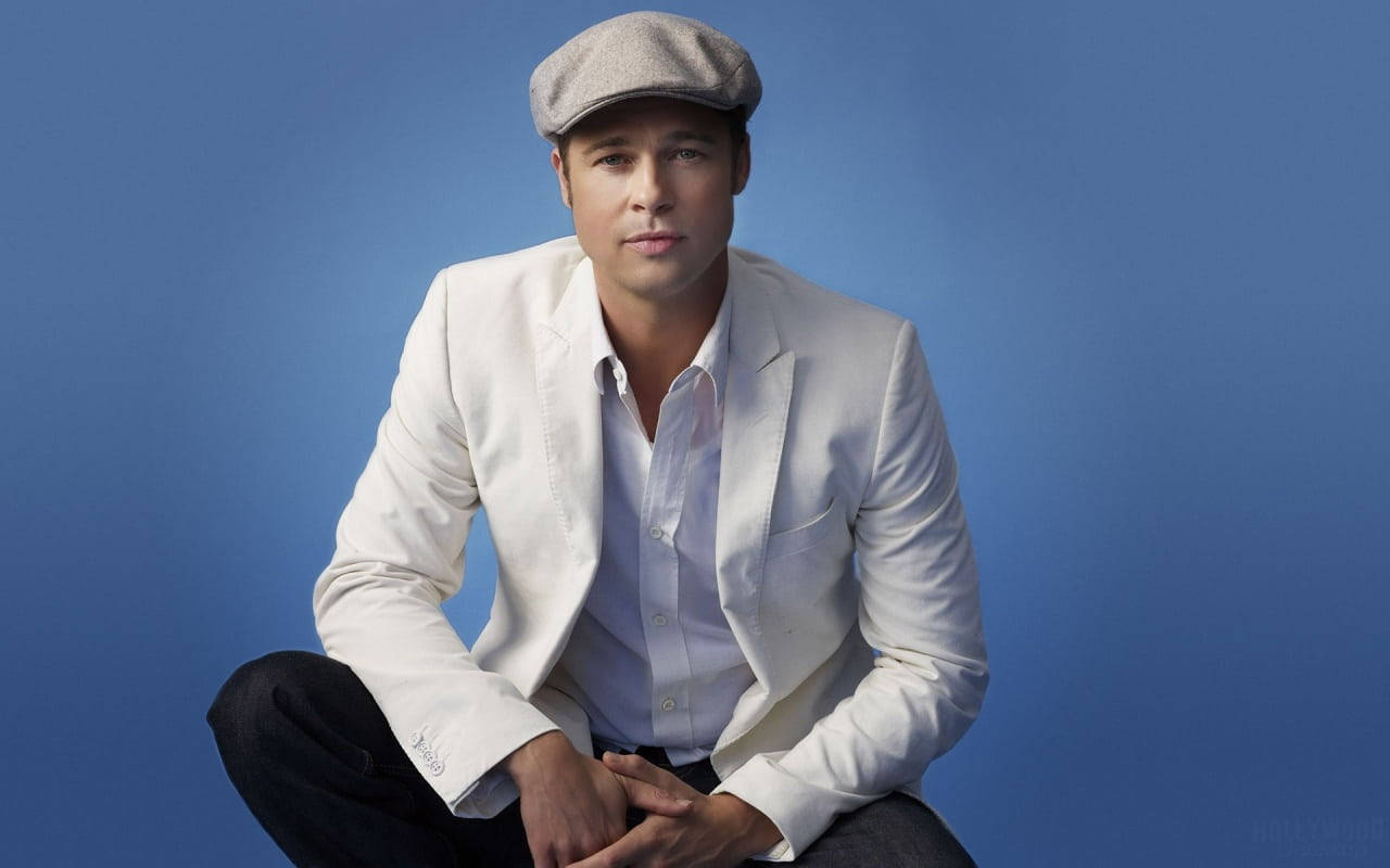 Brad Pitt 桌布 壁紙 — 一名身穿白色西裝外套和灰色帽子的男子自信地坐在純藍色攝影棚背景前。