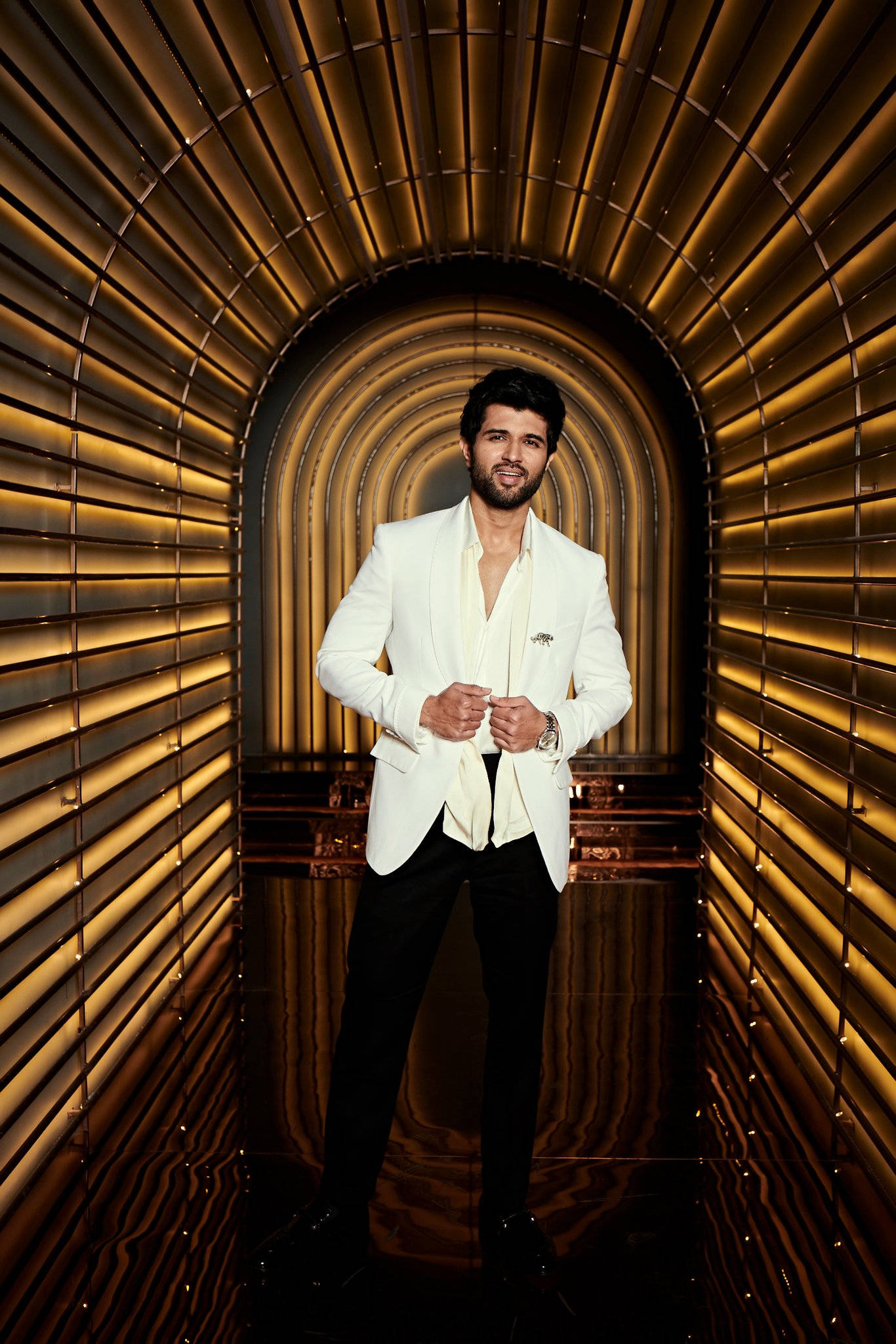 vijay HD 桌布 壁紙 — 一名身穿白色西裝的男子自信地站在一個帶有發光垂直面板的金色拱形走廊中。