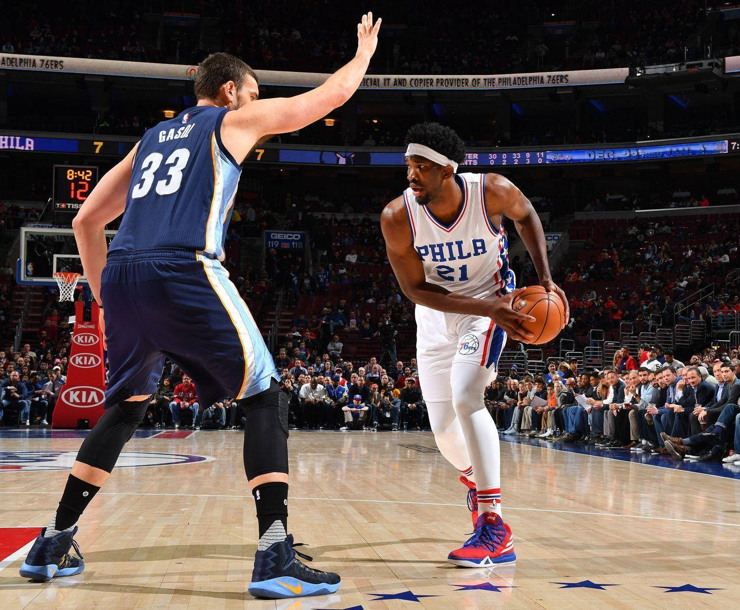 Embiid 桌布 壁紙 — 一名身穿白色費城球衣的籃球球員在比賽中運球，同時被一名身穿藍色球衣的對手防守。