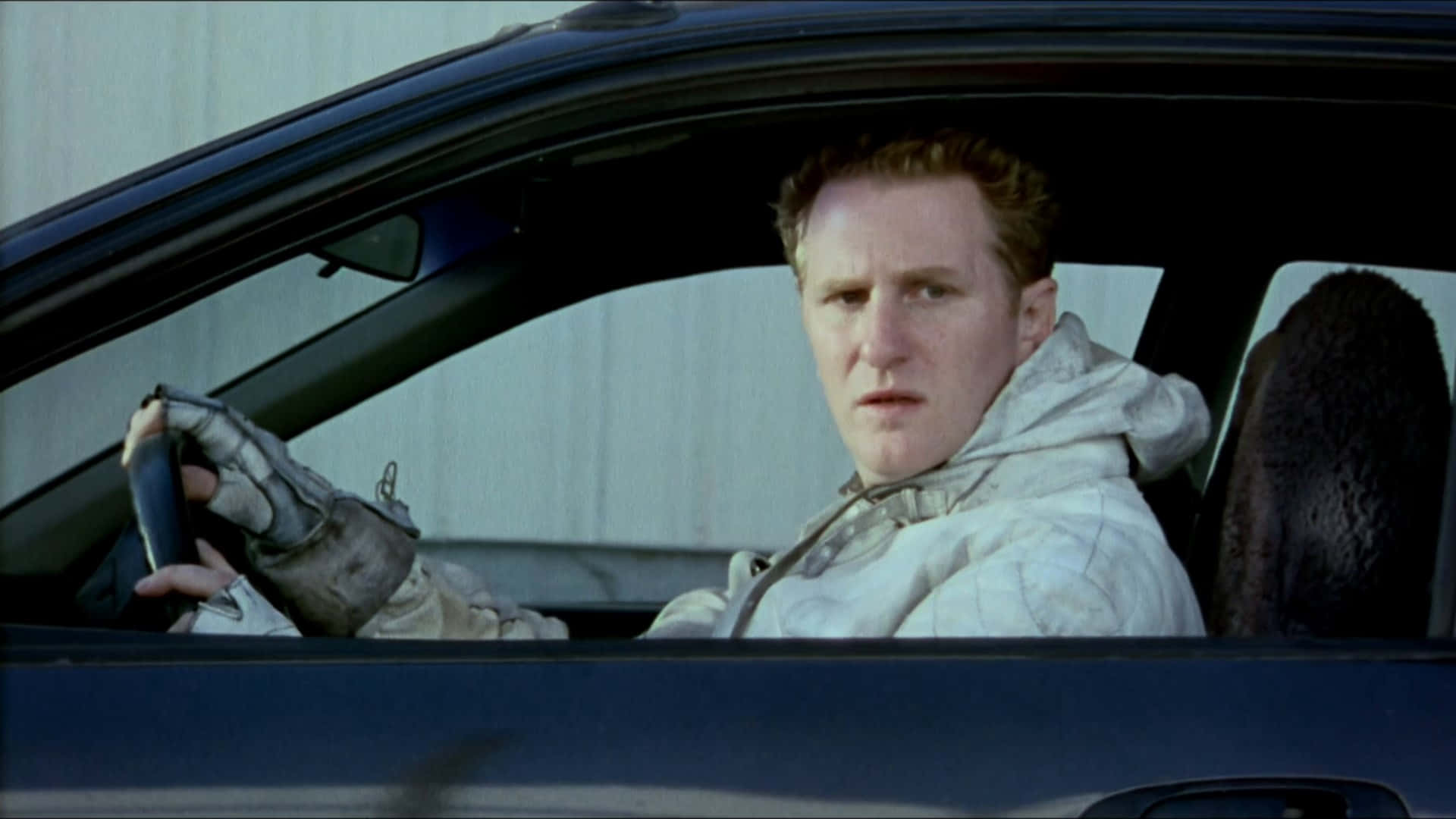 Michael Rapaport 桌布 壁紙 — 一名身穿白色賽車服和手套的男子在黑暗的車內緊握方向盤，在純色牆壁背景下顯得緊張。