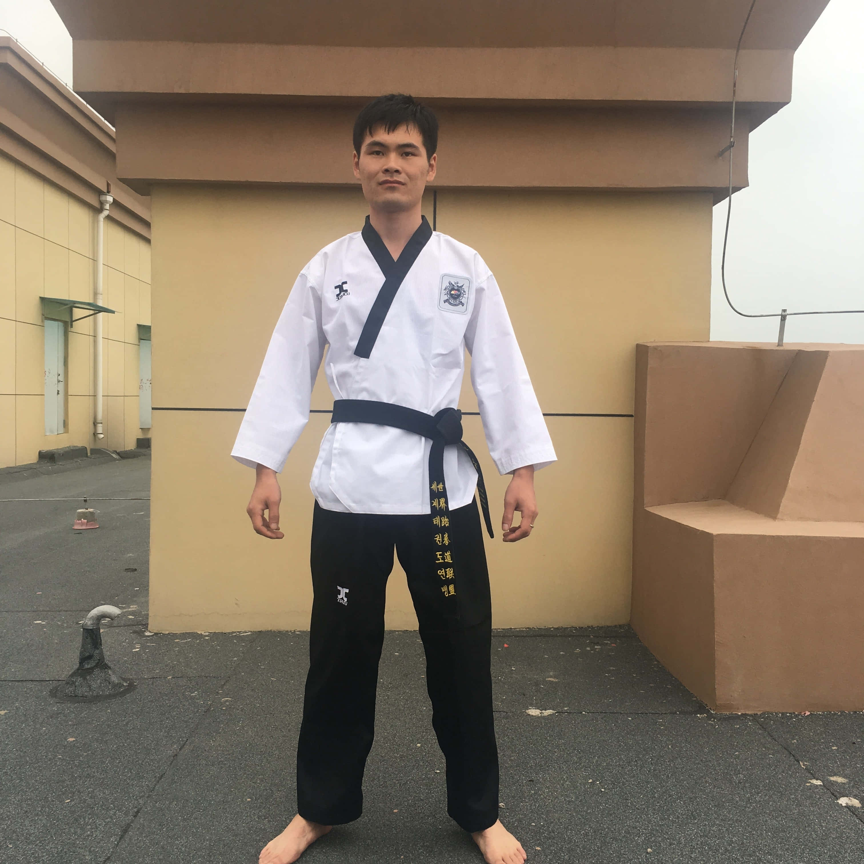 Taekwondo Uniform 桌布 壁紙 — 一名身穿白色跆拳道服、繫著黑帶的年輕男子自信地赤腳站在屋頂上，背景是米色建築牆壁。