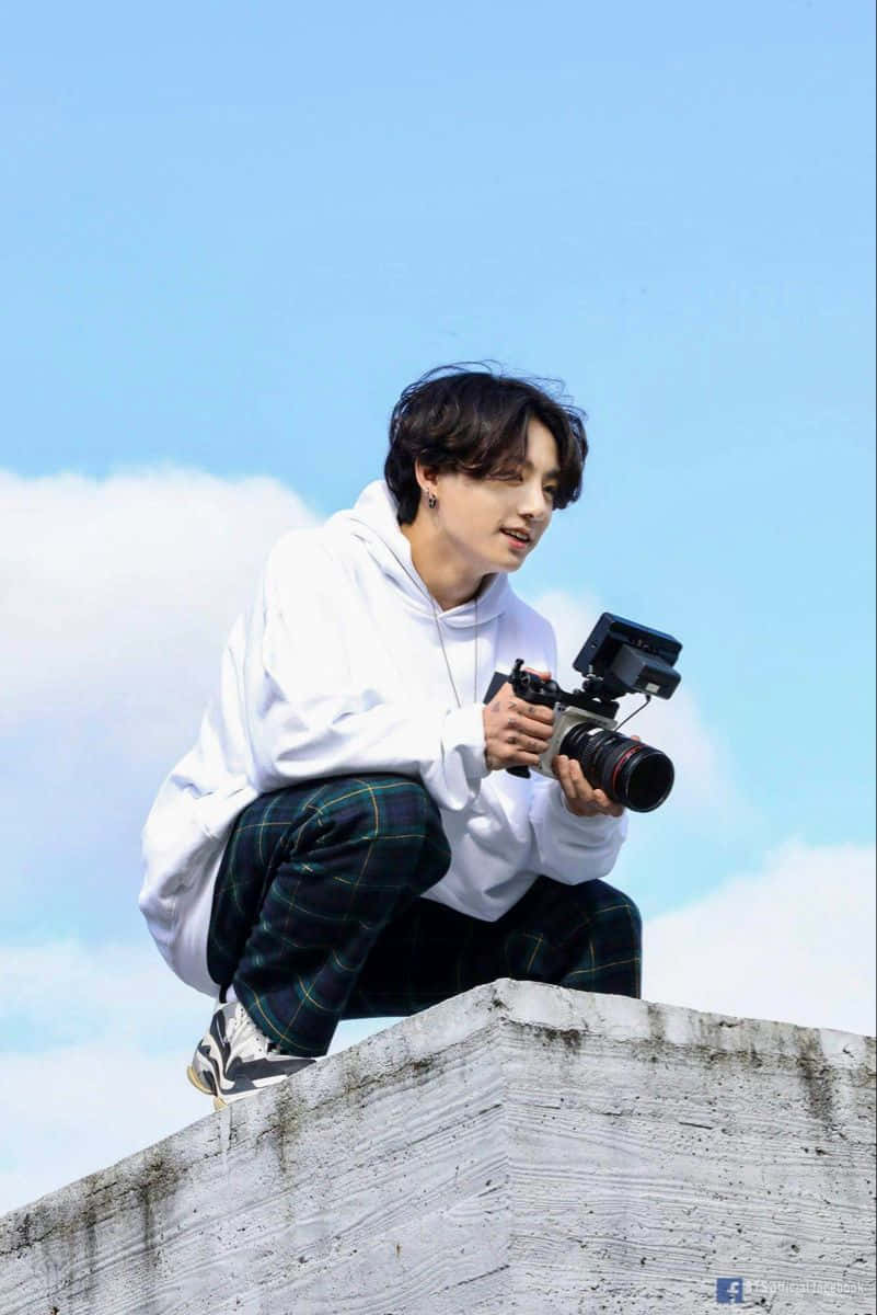 Holding Camera 桌布 壁紙 — 一名身穿白色連帽衫和格子褲的年輕男子蹲在混凝土邊緣，手持相機，背景是藍天白雲。