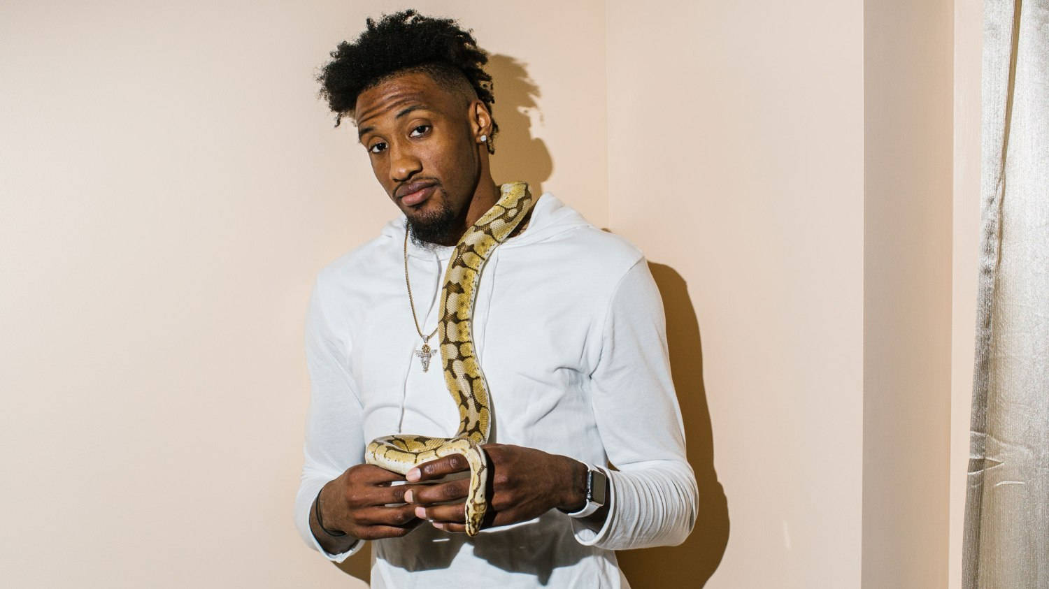 robert covington 桌布 壁紙 — 一名身穿白色連帽衫的男子，在柔和燈光下，手持一條帶有圖案的蛇，背景是純色米色牆壁。