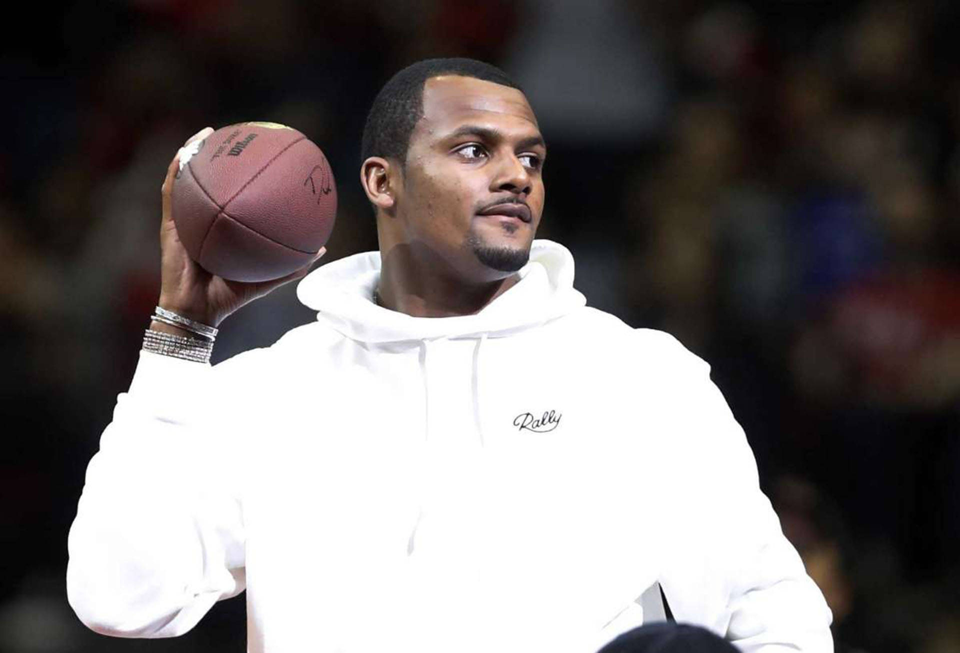 Deshaun Watson 桌布 壁紙 — 一名身穿白色連帽衫的男子，在模糊的體育場人群中，專注地高舉著一個美式足球。
