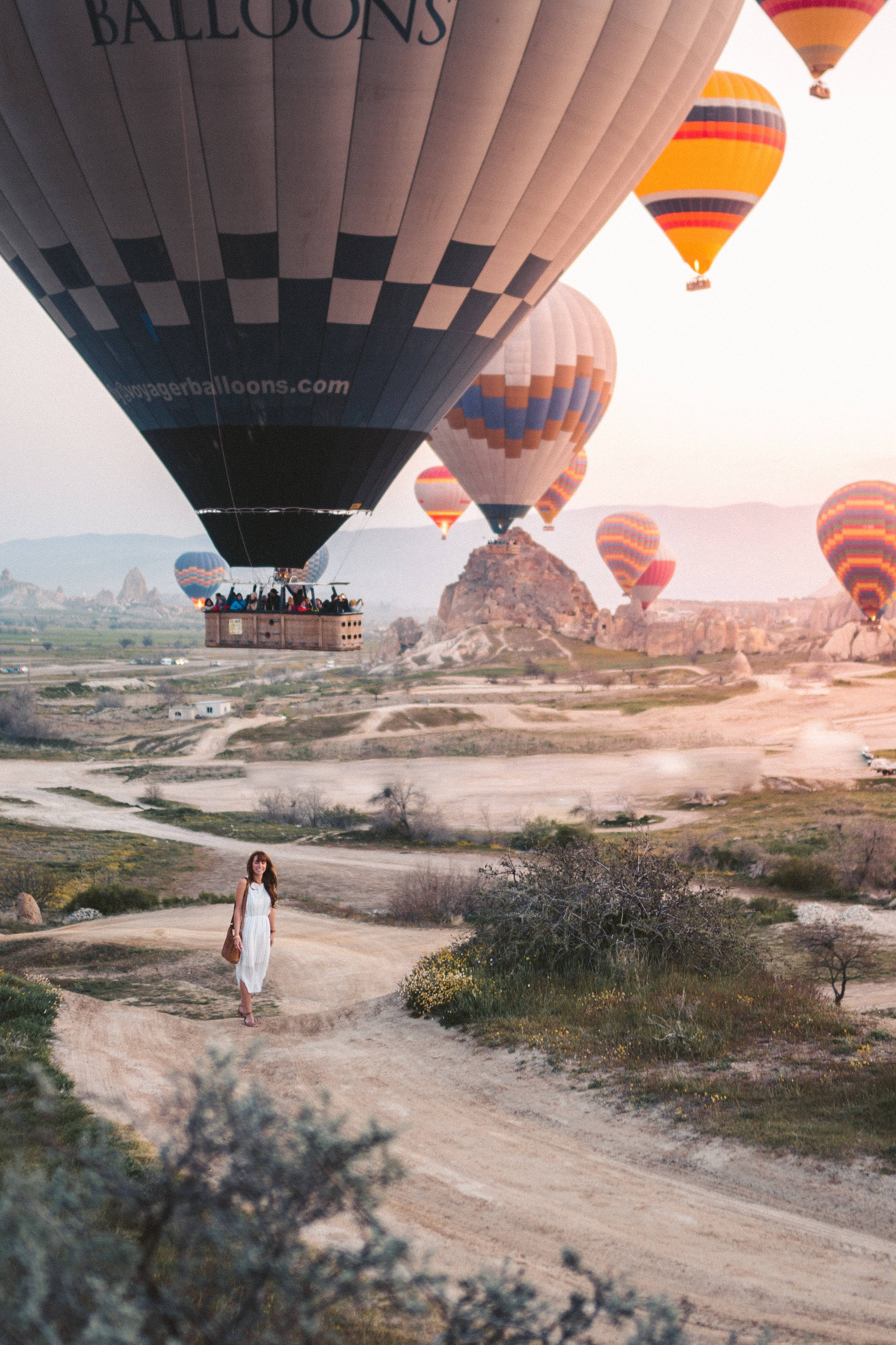 Cappadocia 桌布 壁紙 — 一名身穿白色連衣裙的女子在日出時分，在崎嶇山谷上空飄浮的彩色熱氣球下的泥土小徑上行走。