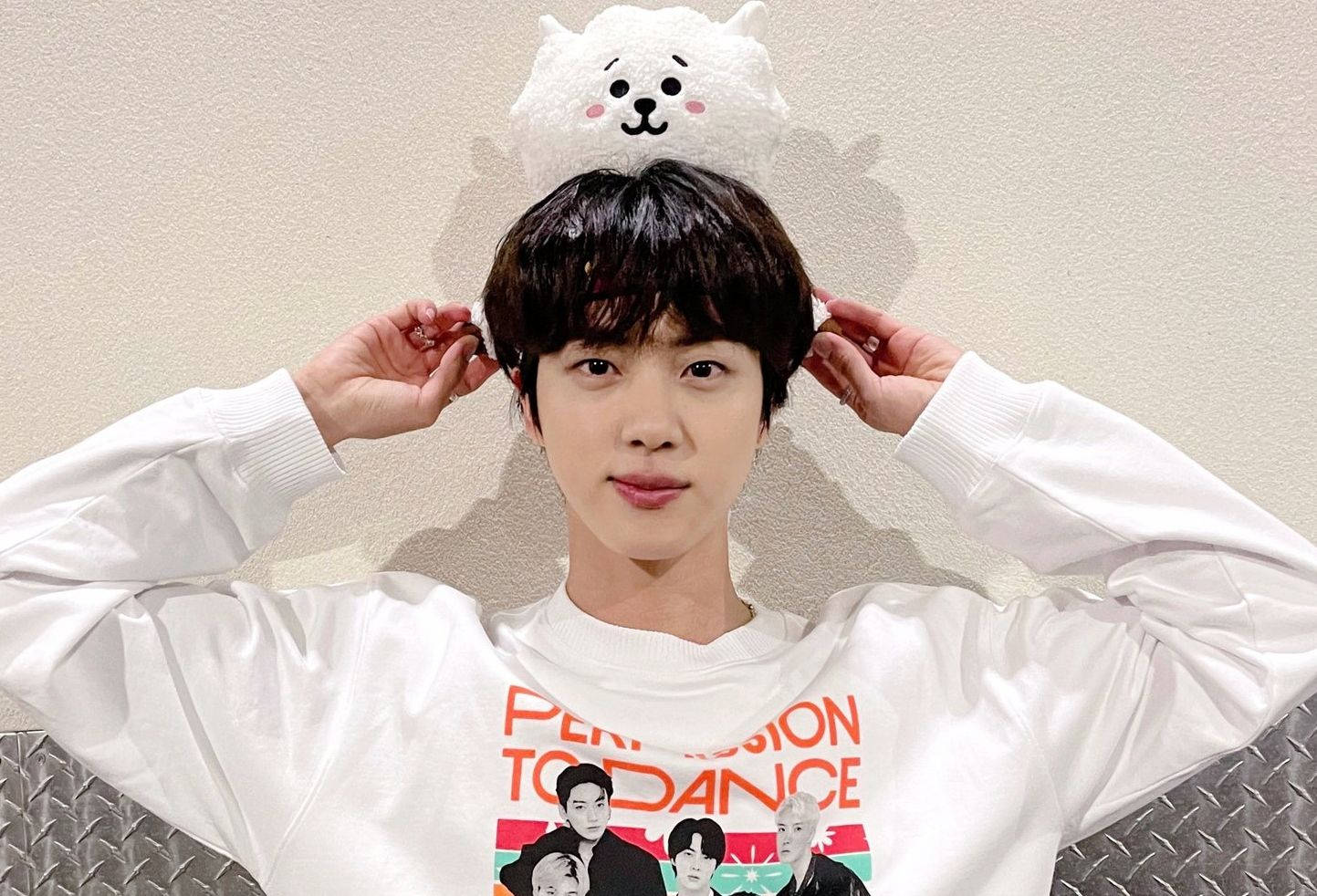 kim seok jin 桌布 壁紙 — 一名身穿白色運動衫的年輕男子在室內頑皮地將一個毛茸茸的白色貓咪玩偶平衡在頭上。