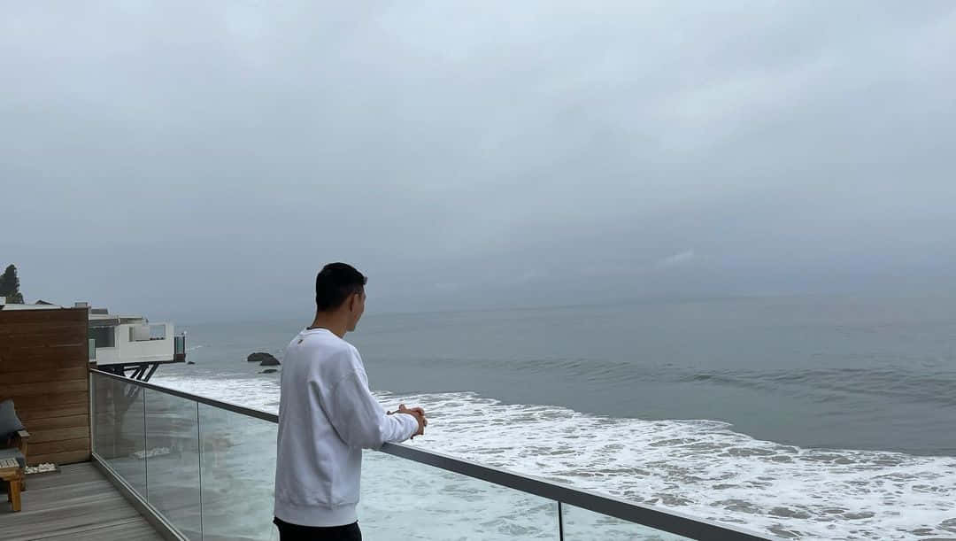 海灘 桌布 壁紙 — 一名身穿白色運動衫的男子站在玻璃欄杆的陽台上，俯瞰著陰天籠罩下霧氣瀰漫的海洋。
