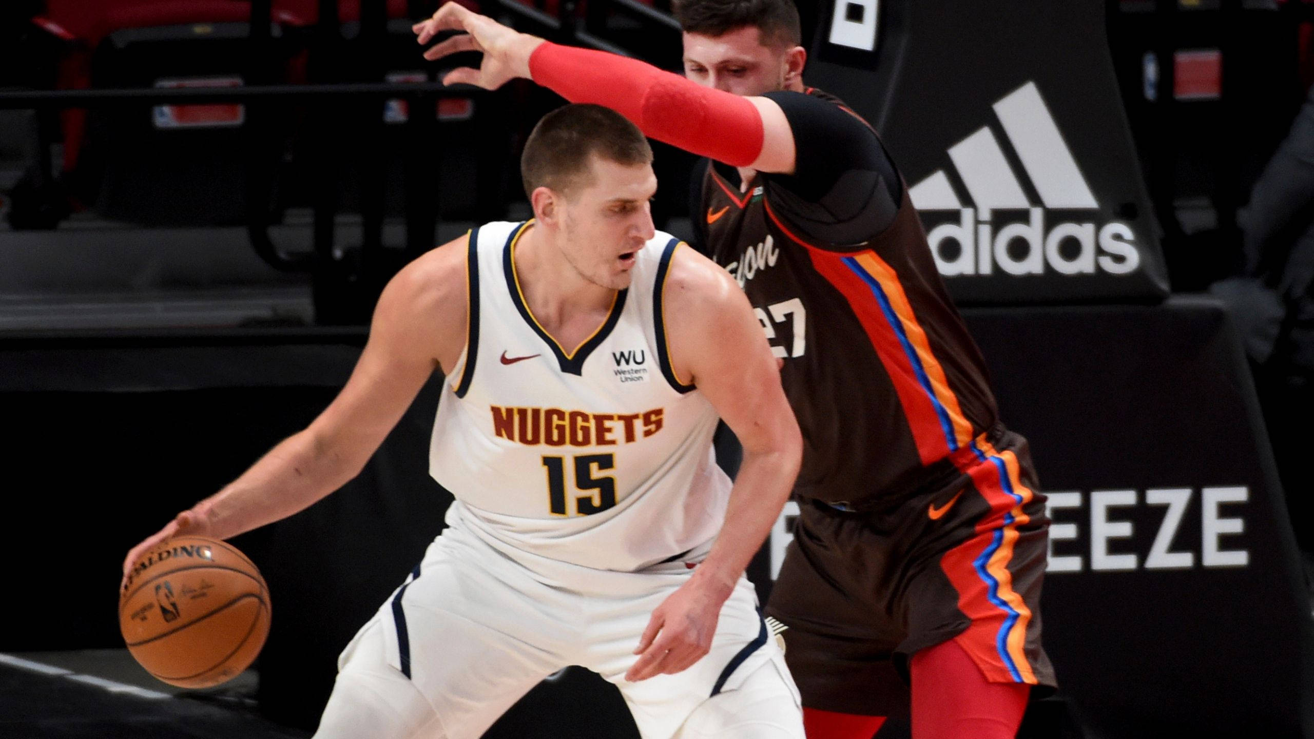 Jokic 桌布 壁紙 — 一名身穿白色金塊球衣的籃球運動員在激烈的比賽中運球，同時被一名身穿棕色球衣的對手緊密防守。