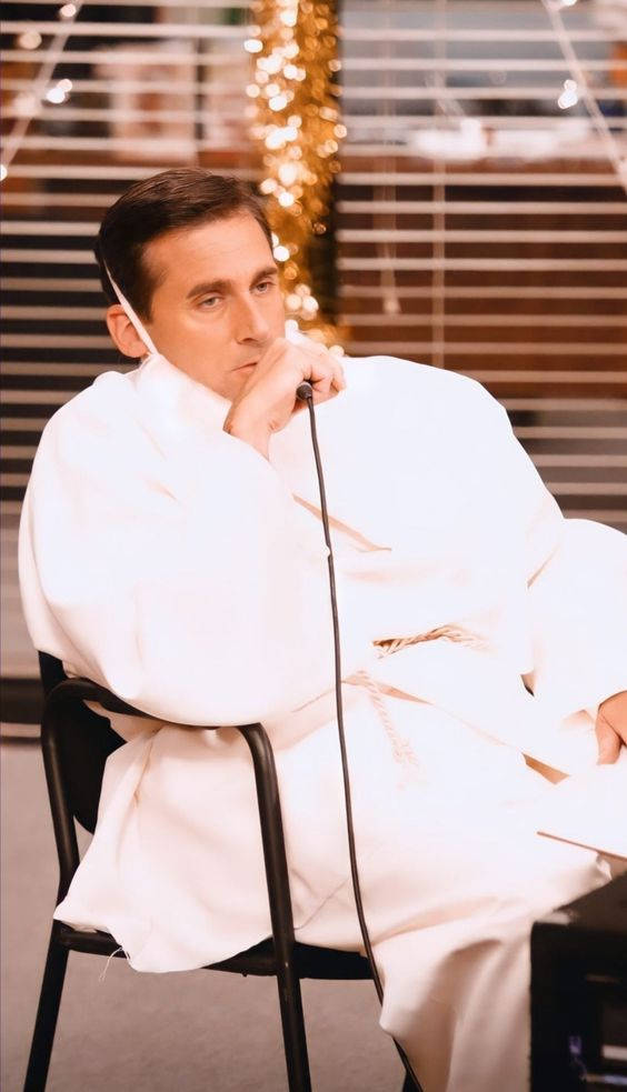 Michael Scott 桌布 壁紙 — 一名身穿白色長袍的男子坐在椅子上，嘴邊放著麥克風，沉思著，周圍環繞著溫暖的串燈和百葉窗。