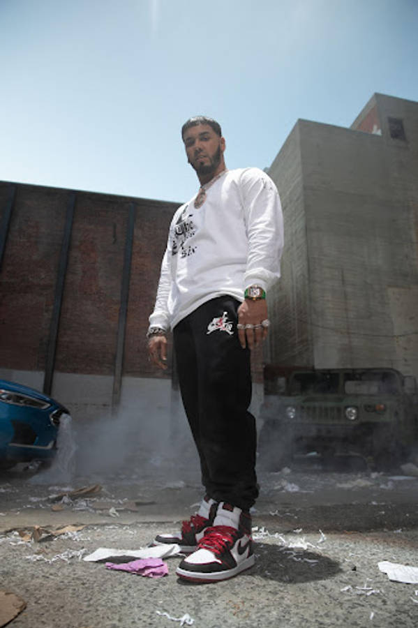 Anuel Aa 桌布 壁紙 — 一名身穿白色長袖衫和黑色褲子的男子自信地站在城市小巷中，煙霧在他腳邊升起。