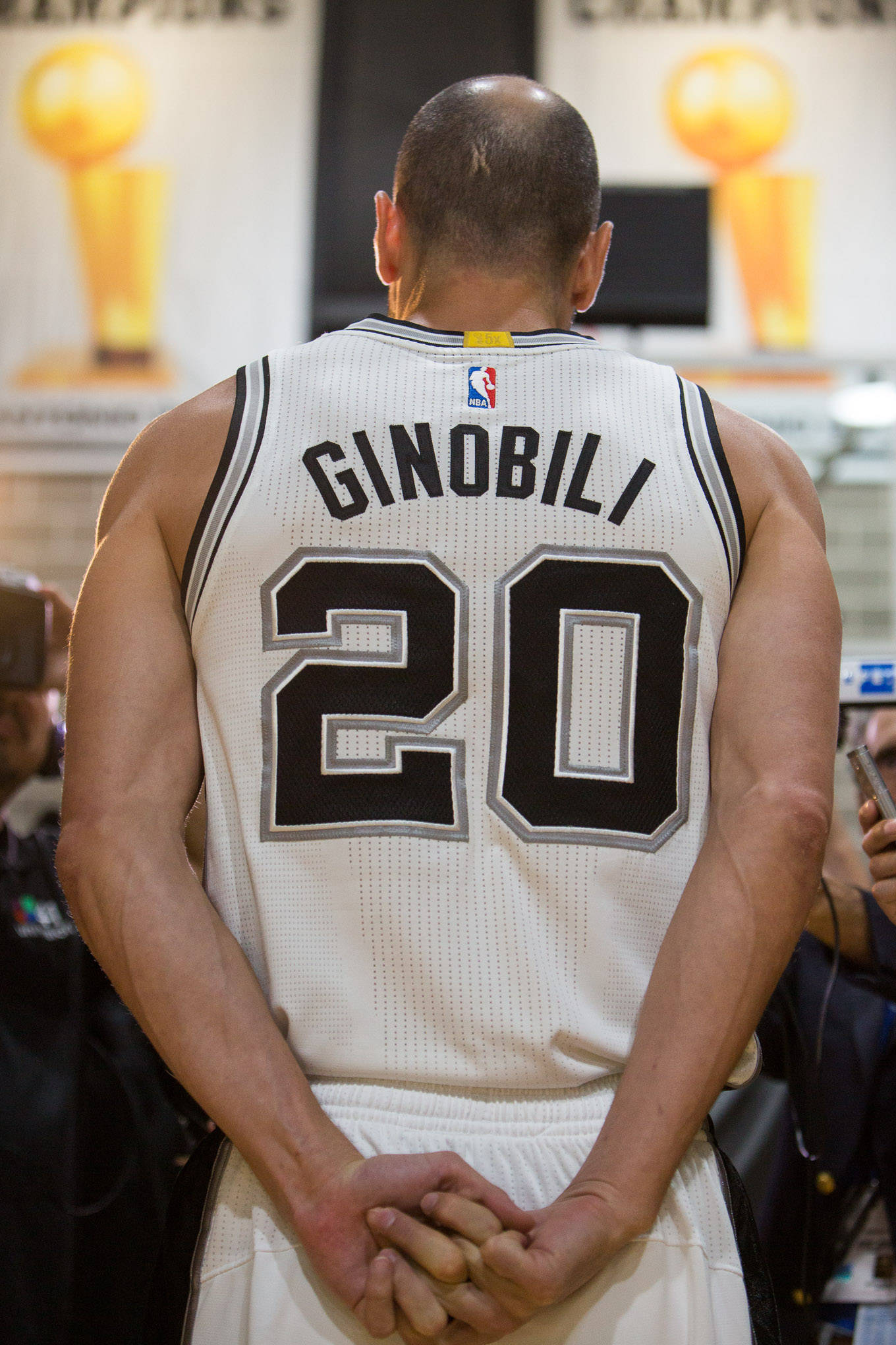 Manu 桌布 壁紙 — 一名身穿白色馬刺球衣、背號20的籃球運動員雙手合十，面向兩張冠軍獎盃海報站立。
