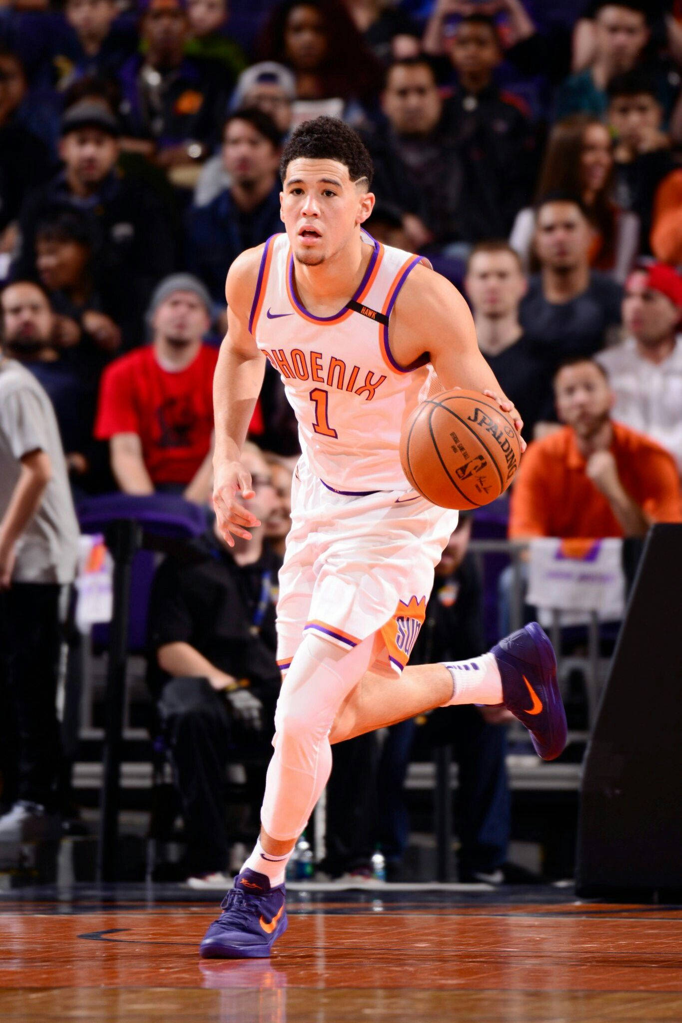 Devin Booker 桌布 壁紙 — 一名身穿白色鳳凰城太陽隊球衣的籃球運動員在激烈的比賽中運球，背景是人群。