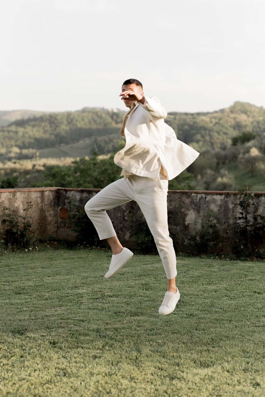 Minimalist Fashion 桌布 壁紙 — 一名身穿白衣的男子在草地上跳起，背景是連綿起伏的綠色山丘。