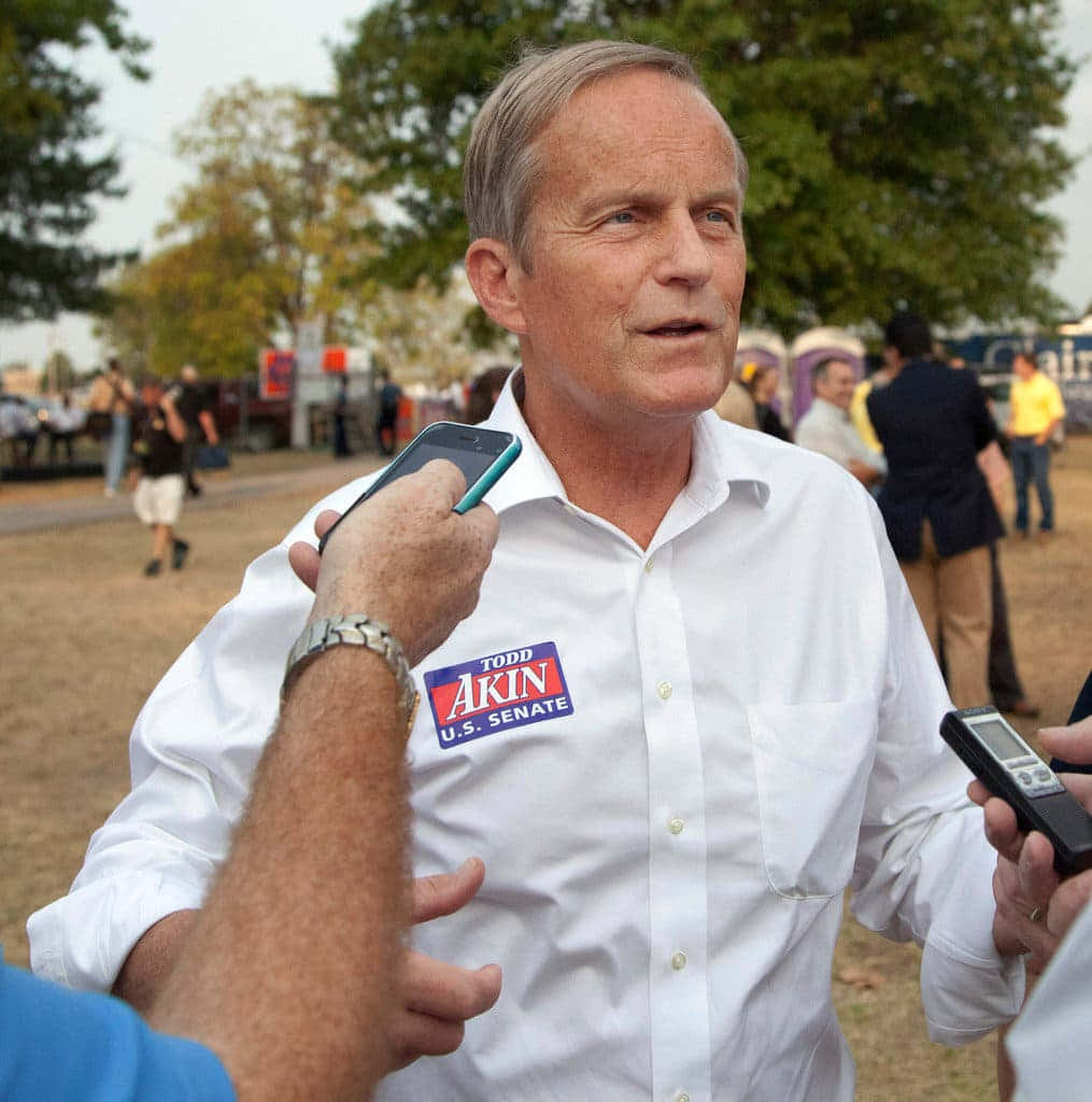 樹木 桌布 壁紙 — 一名身穿白襯衫、佩戴 Todd Akin 美國參議院競選徽章的男子，在白天戶外向記者發言。
