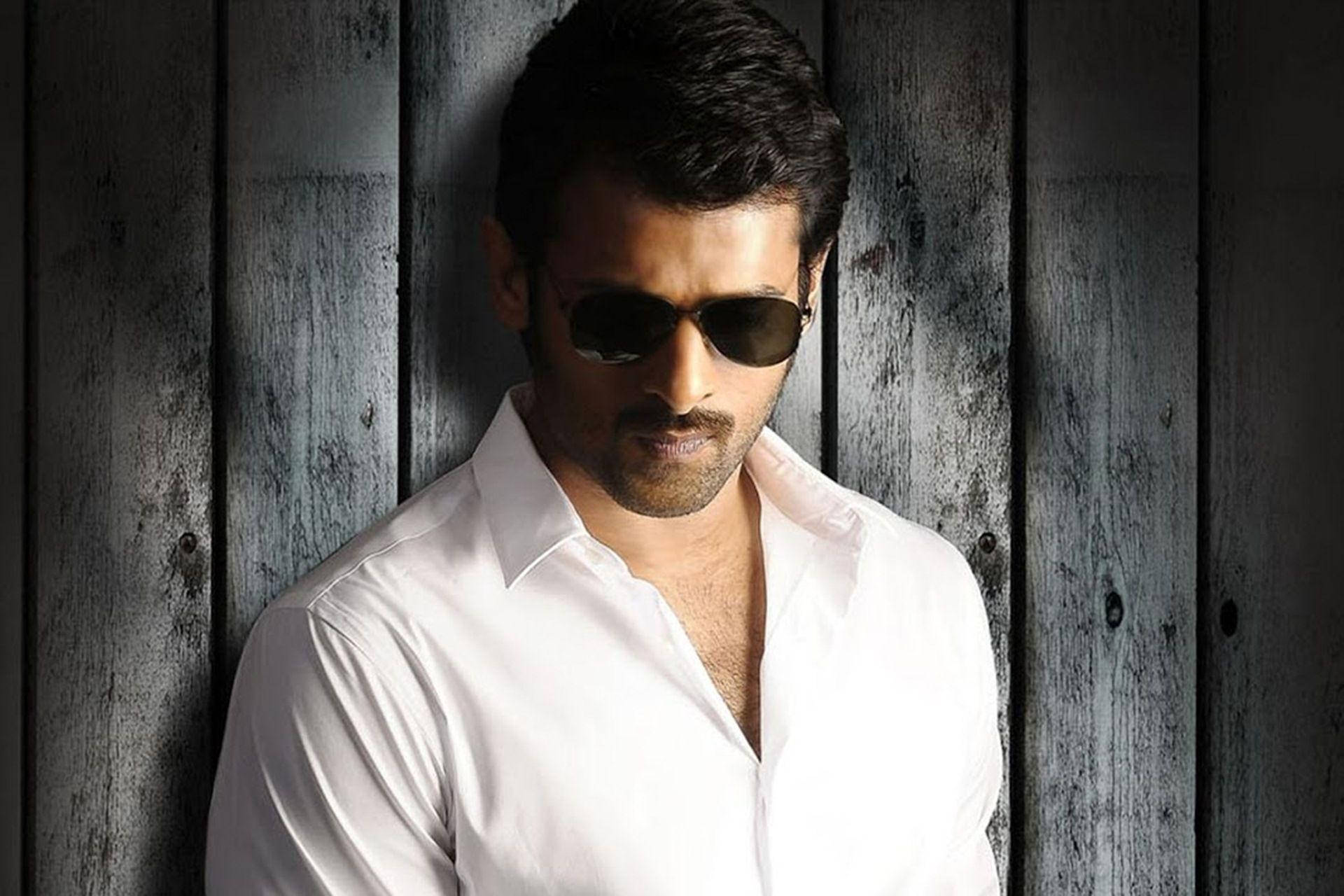Prabhas HD 桌布 壁紙 — 一名身穿白襯衫和太陽眼鏡的男子，在戲劇性的燈光下靠著風化的灰色木板。