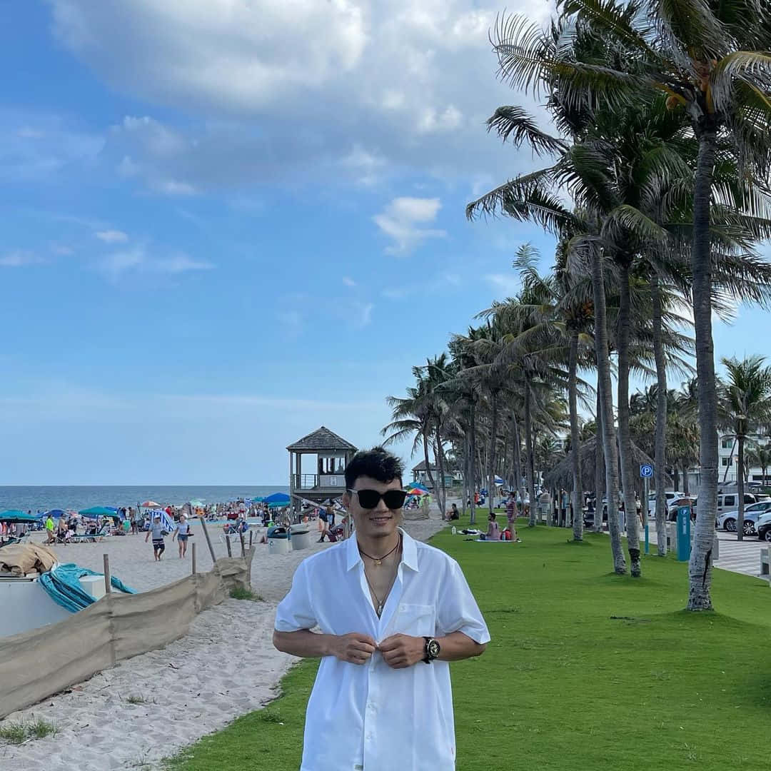 海灘 桌布 壁紙 — 一名身穿白襯衫和太陽眼鏡的男子在陽光明媚、棕櫚樹和沙灘遊客林立的海灘上微笑站立。