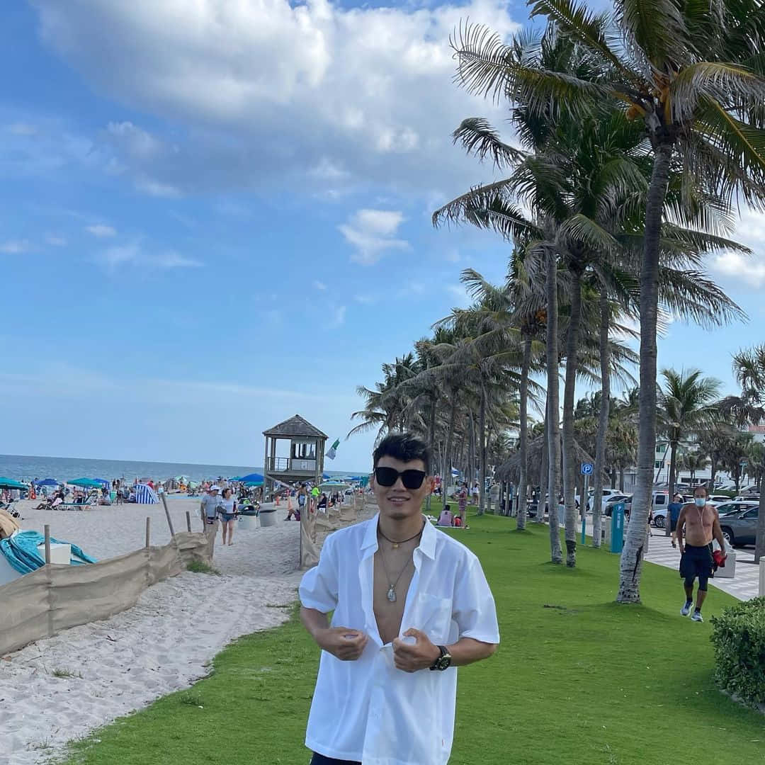 海灘 桌布 壁紙 — 一名身穿白襯衫和太陽眼鏡的男子站在棕櫚樹旁的草地上，背景是繁忙的海灘和救生員瞭望塔，天空部分多雲。