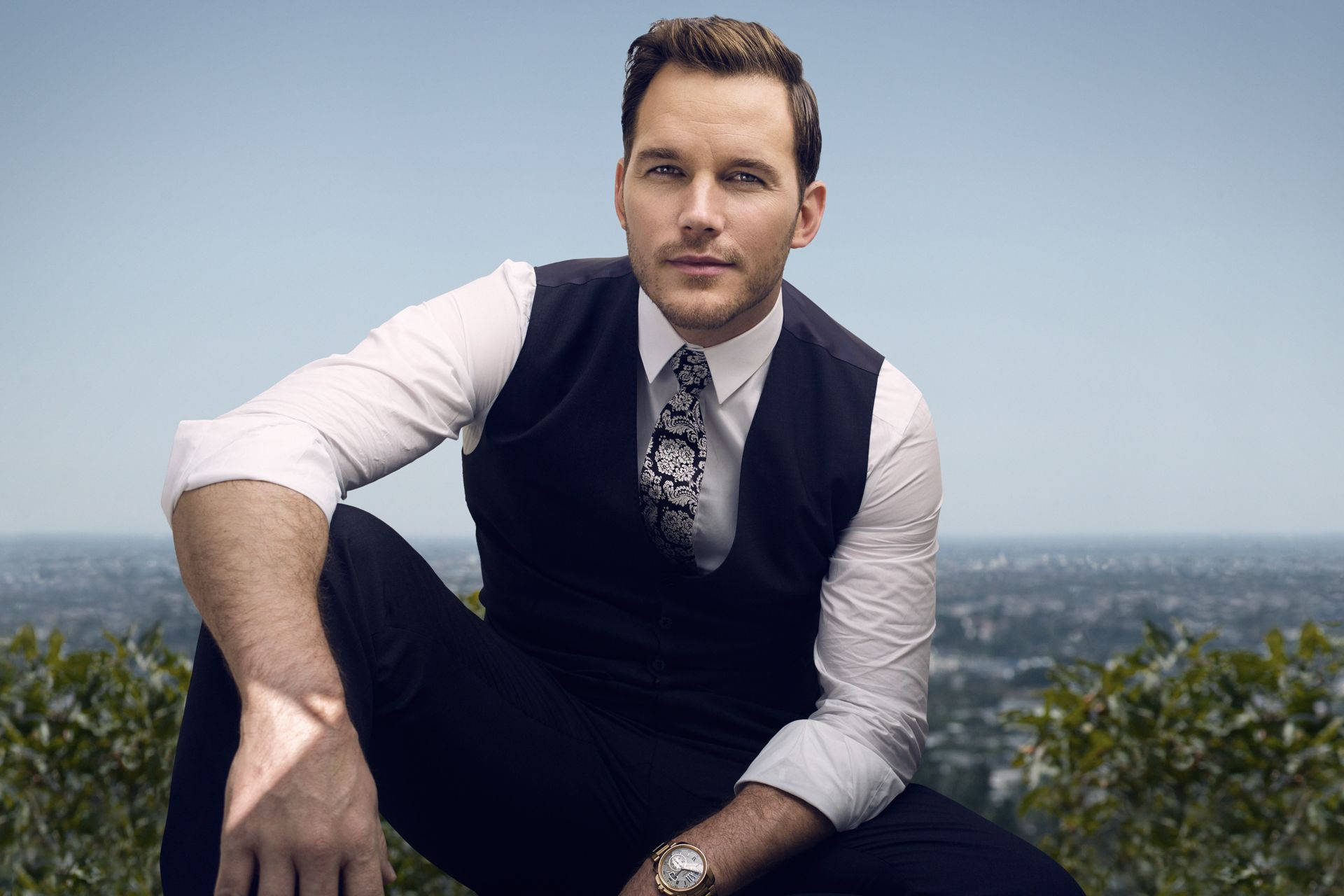 Chris Pratt 桌布 壁紙 — 一名身穿白襯衫和深色背心的男子自信地坐在山頂上，俯瞰著晴朗藍天下廣闊的城市景觀。
