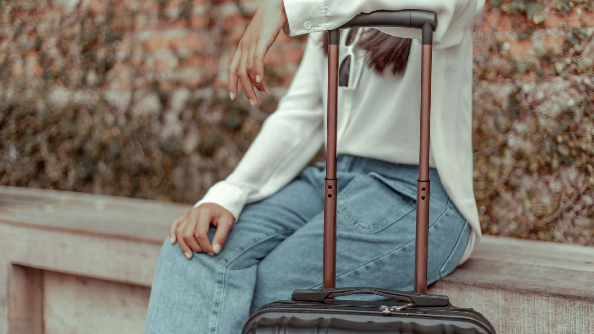 Beautiful Travel 桌布 壁紙 — 一名身穿白襯衫和牛仔褲的人坐在木長椅上，旁邊放著一個帶輪行李箱，背景是磚牆。