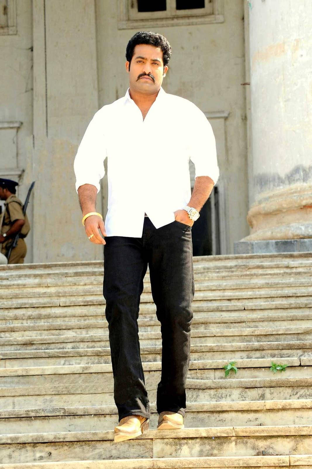 jr ntr 桌布 壁紙 — 一名身穿白襯衫和黑褲的男子自信地站在古典建築前的寬闊石階上。