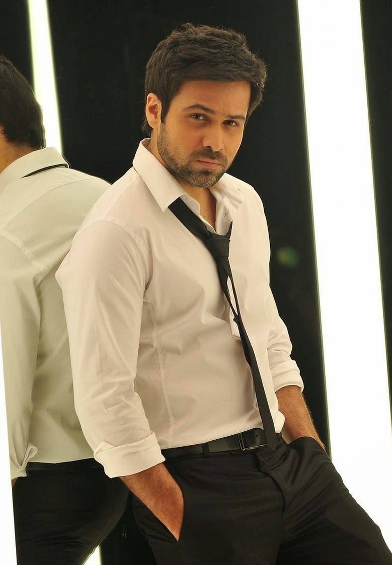 Emraan Hashmi 桌布 壁紙 — 一名身穿白襯衫和黑領帶的男子，雙手插袋，在黑白條紋背景前自信地擺姿勢。