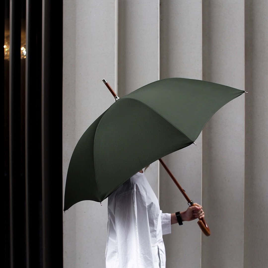 Umbrella 桌布 壁紙 — 一名身穿白襯衫的人拿著一把大型橄欖綠色雨傘，在垂直建築柱子的背景下遮住臉部。