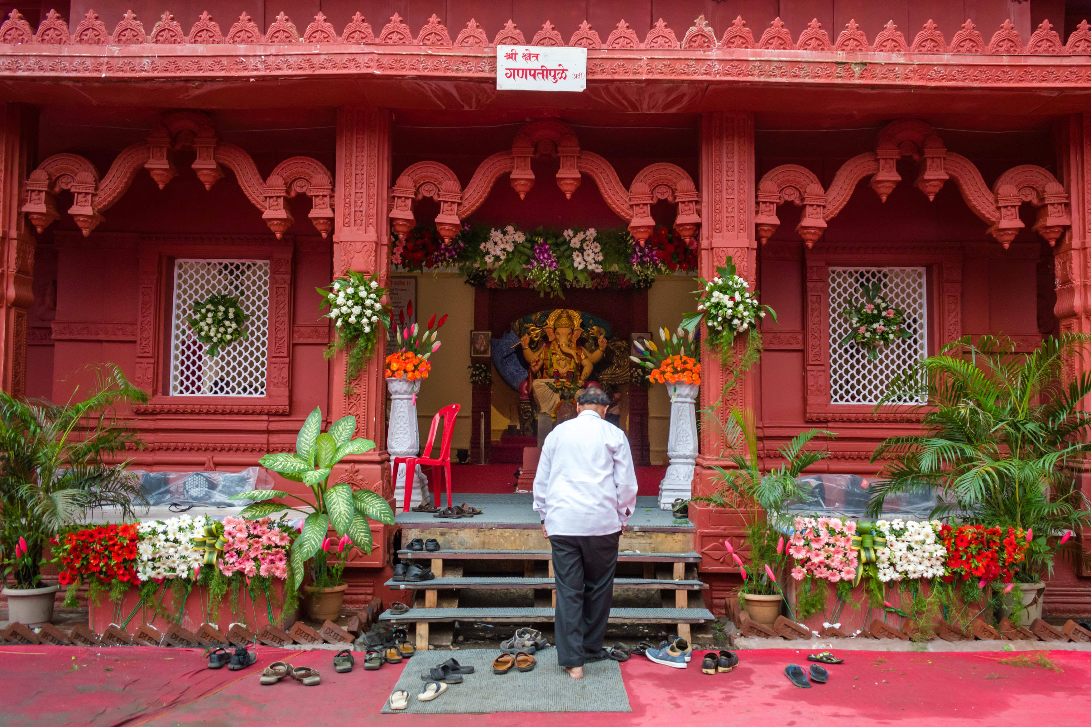 ganesh 4K 桌布 壁紙 — 一名身穿白襯衫的人赤腳站在階梯上，面前是一座以鮮花和華麗雕刻裝飾的鮮紅色印度教寺廟。