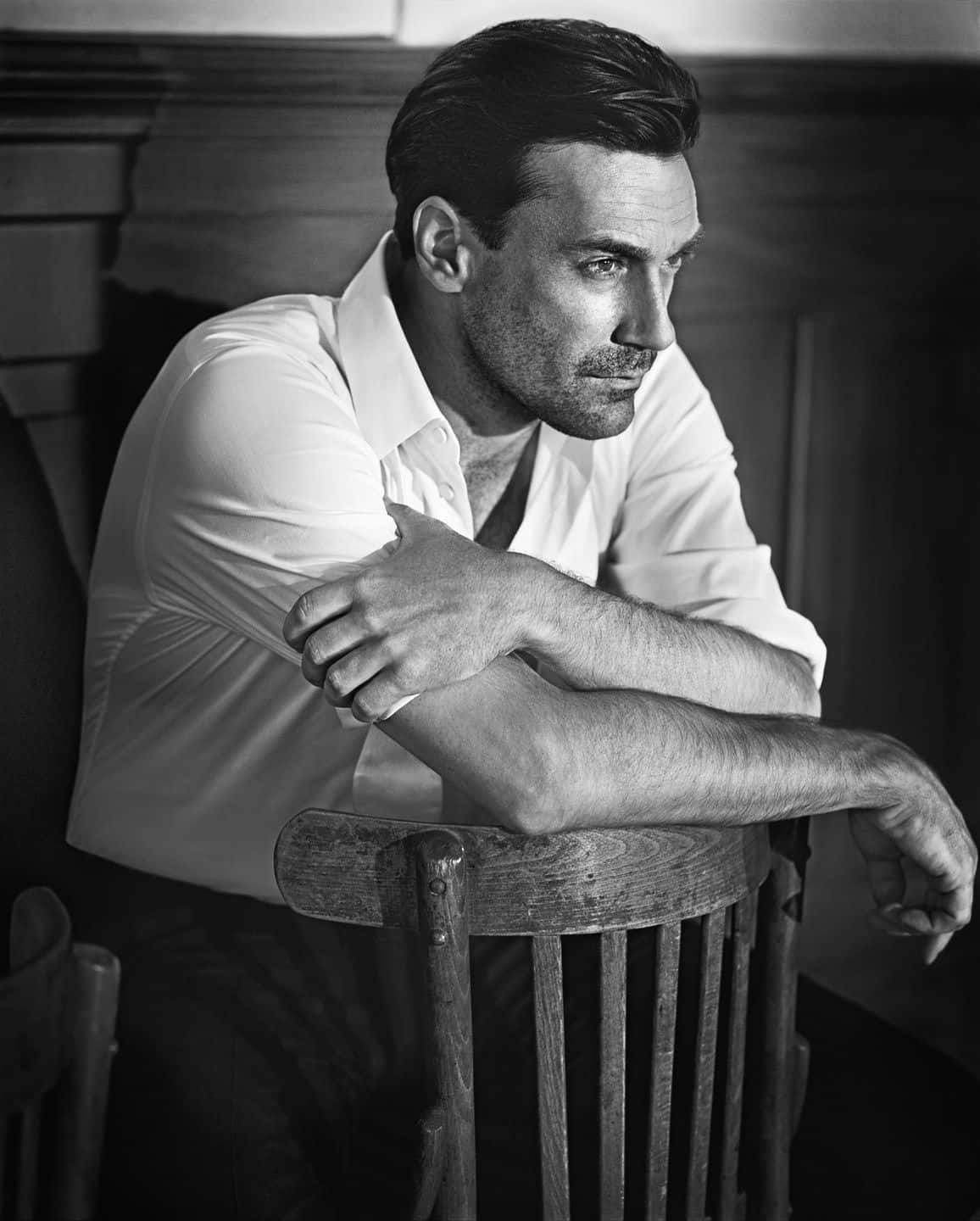 Jon Hamm 桌布 壁紙 — 一名身穿白襯衫的男子在昏暗的房間裡若有所思地靠在一張木椅上，以引人注目的黑白影像捕捉。