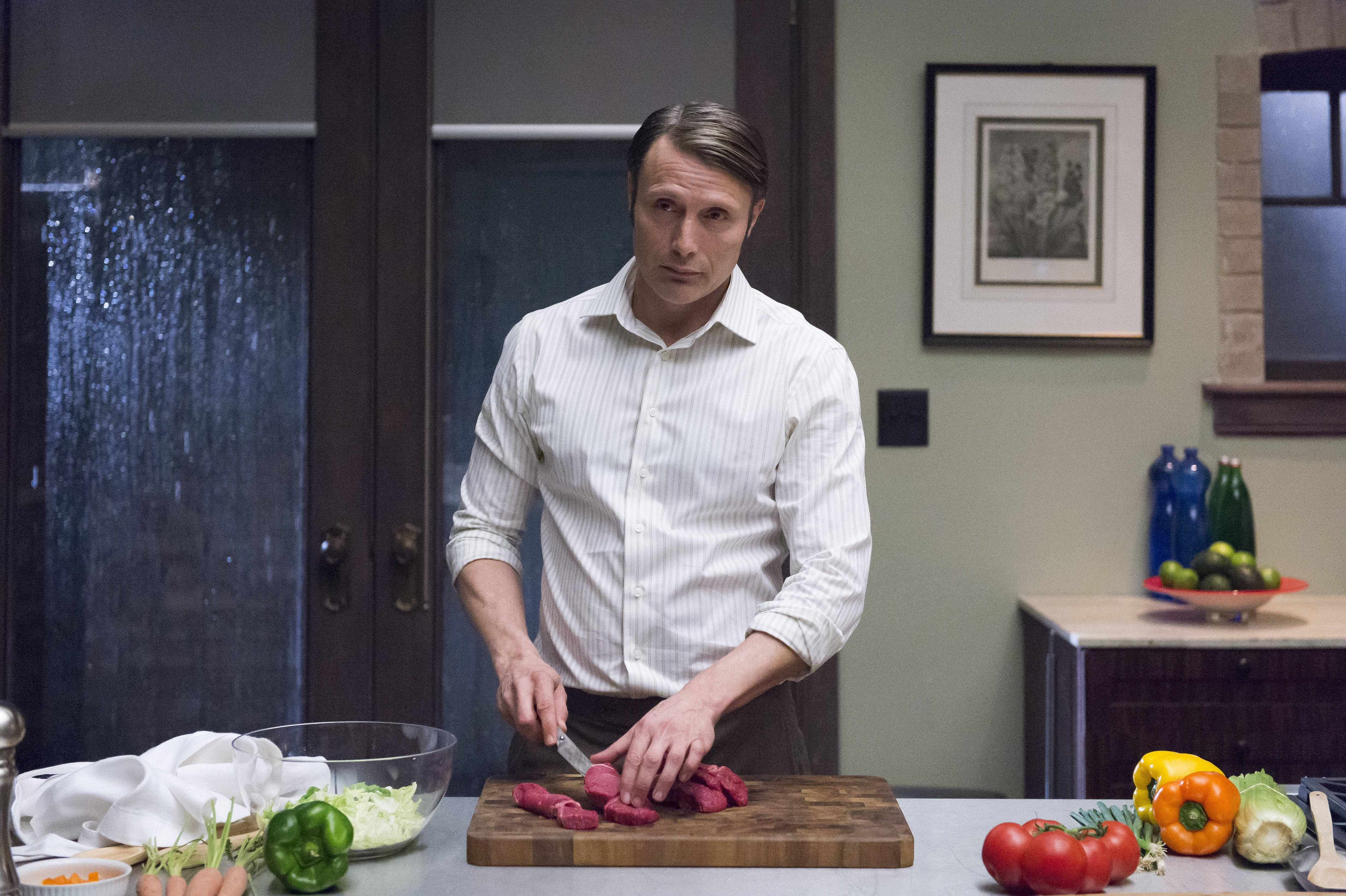 Mads Mikkelsen 桌布 壁紙 — 一名身穿白襯衫的男子在現代化廚房的木製砧板上切生肉，周圍放著新鮮蔬菜。