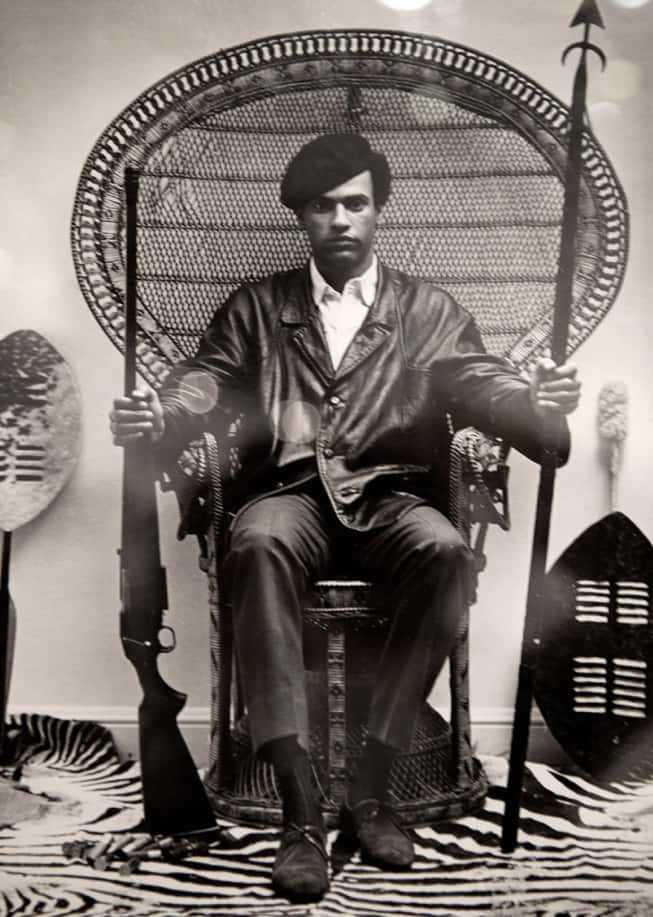 Black Panther Party 桌布 壁紙 — 一名身穿皮夾克和貝雷帽的男子威嚴地坐在華麗的扇形椅上，手持步槍和長矛，兩側有部落盾牌。