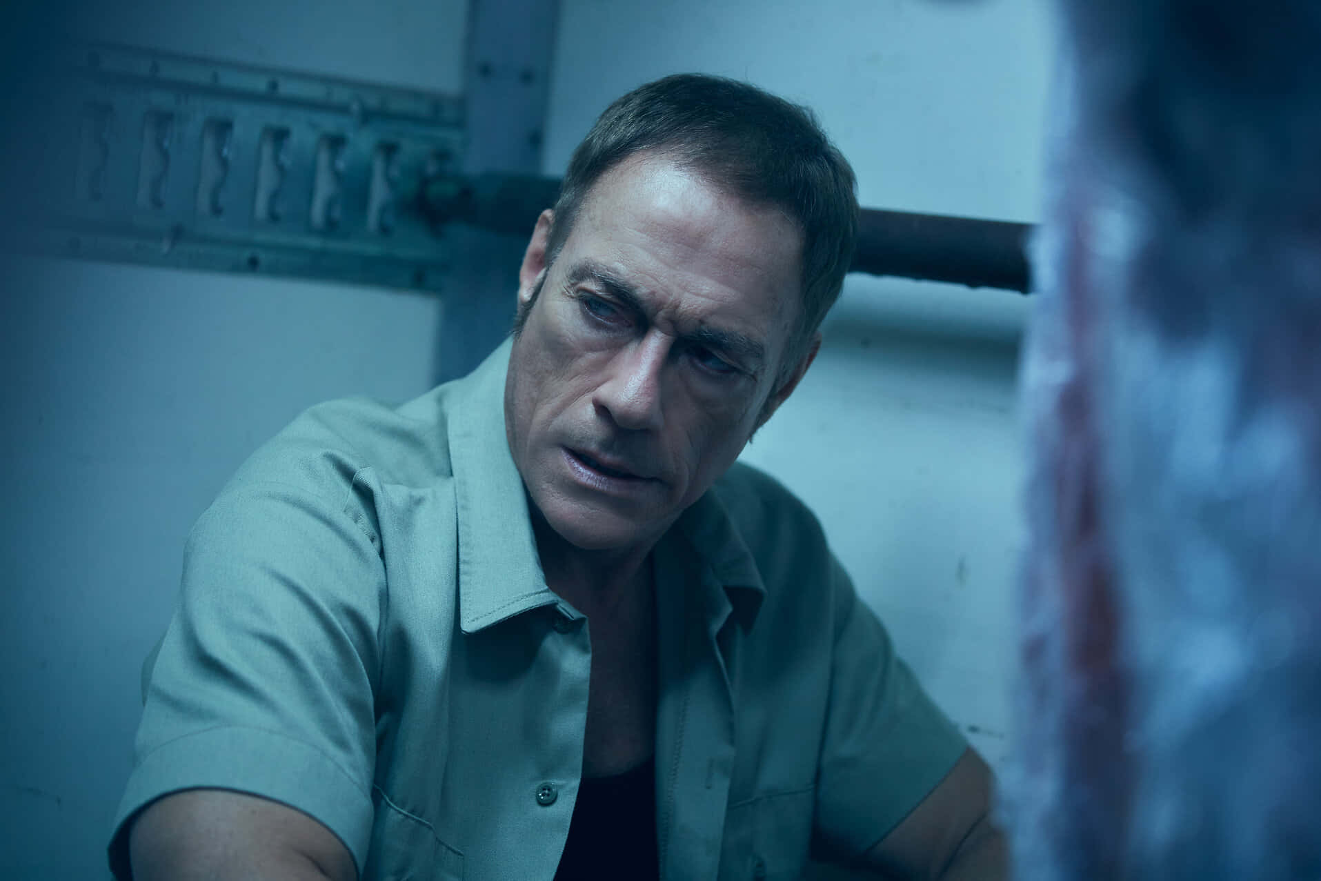 jean claude van damme 桌布 壁紙 — 一名身穿監獄制服的男人坐在昏暗藍光的牢房中，專注地凝視，表情緊張。