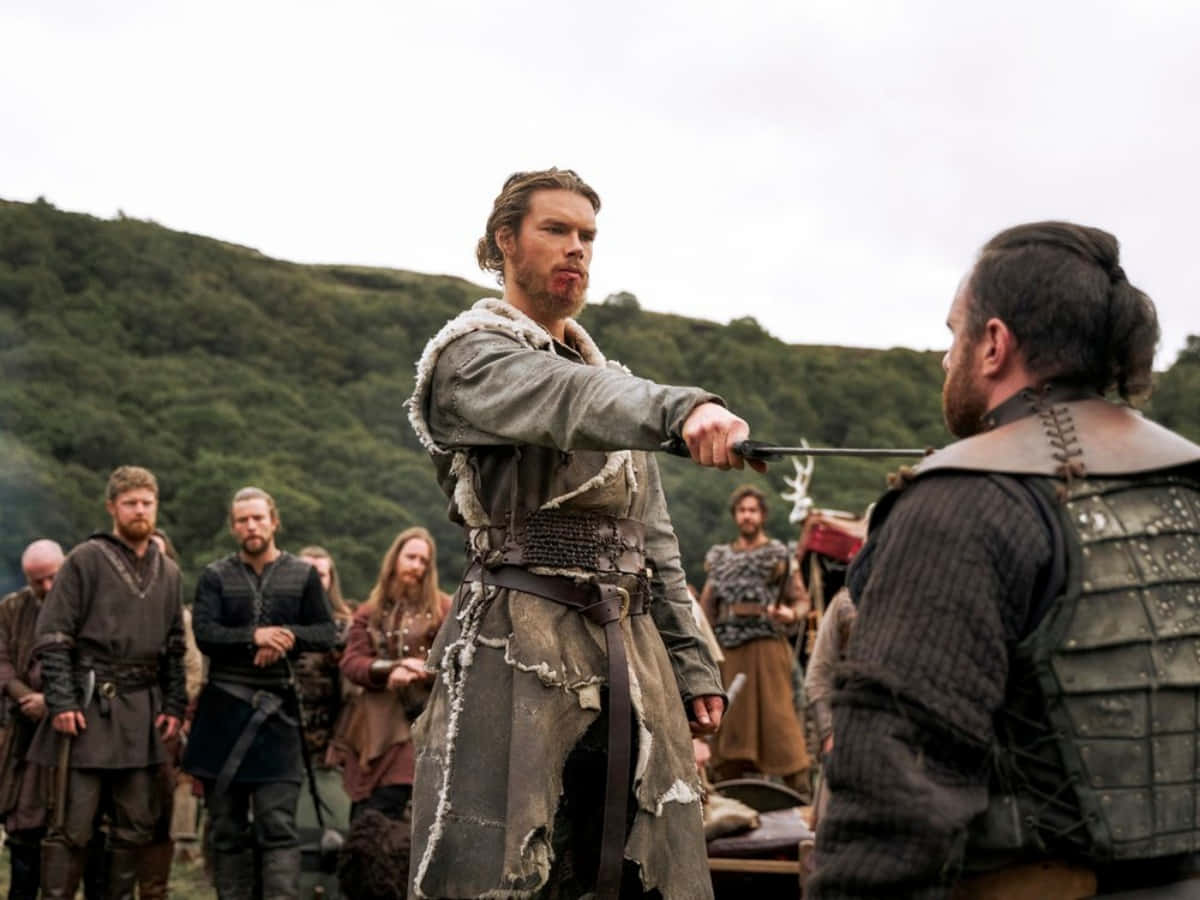 Vikings 桌布 壁紙 — 一名身穿破爛盔甲的維京戰士，在霧氣瀰漫的林間空地中，於旁觀者面前，緊張地持劍刺向對手。