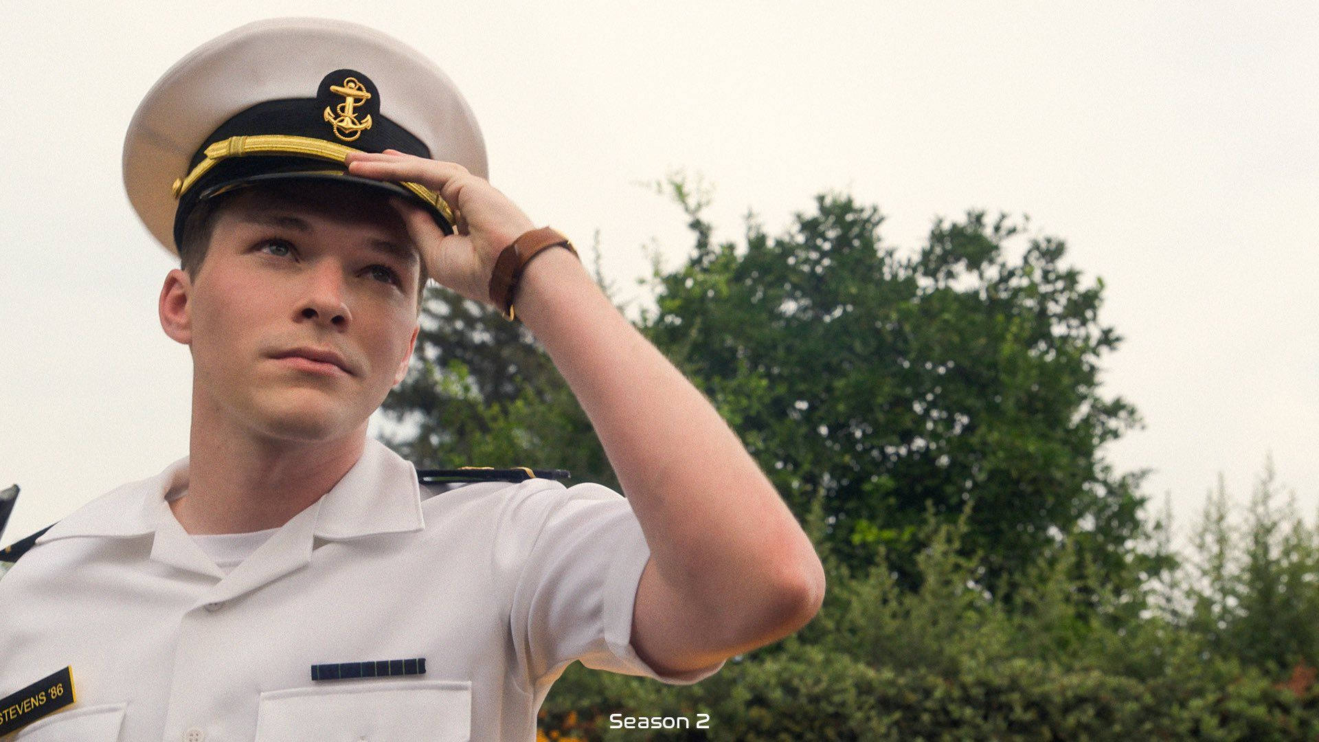 全人類 桌布 壁紙 — 一名身穿筆挺白色制服的年輕海軍軍官，在多雲的天空下，以茂密的綠樹為背景，莊嚴敬禮。
