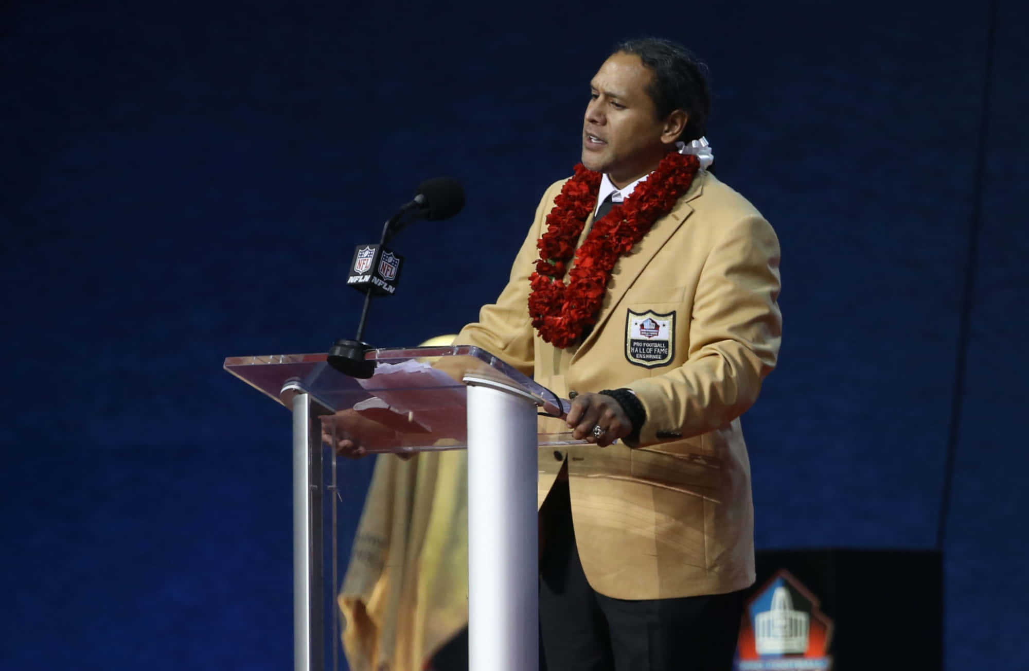 Troy Polamalu 桌布 壁紙 — 一名身穿米色NFL名人堂外套、飾有紅色花環的男子，在深藍色背景前的透明講台上發言。