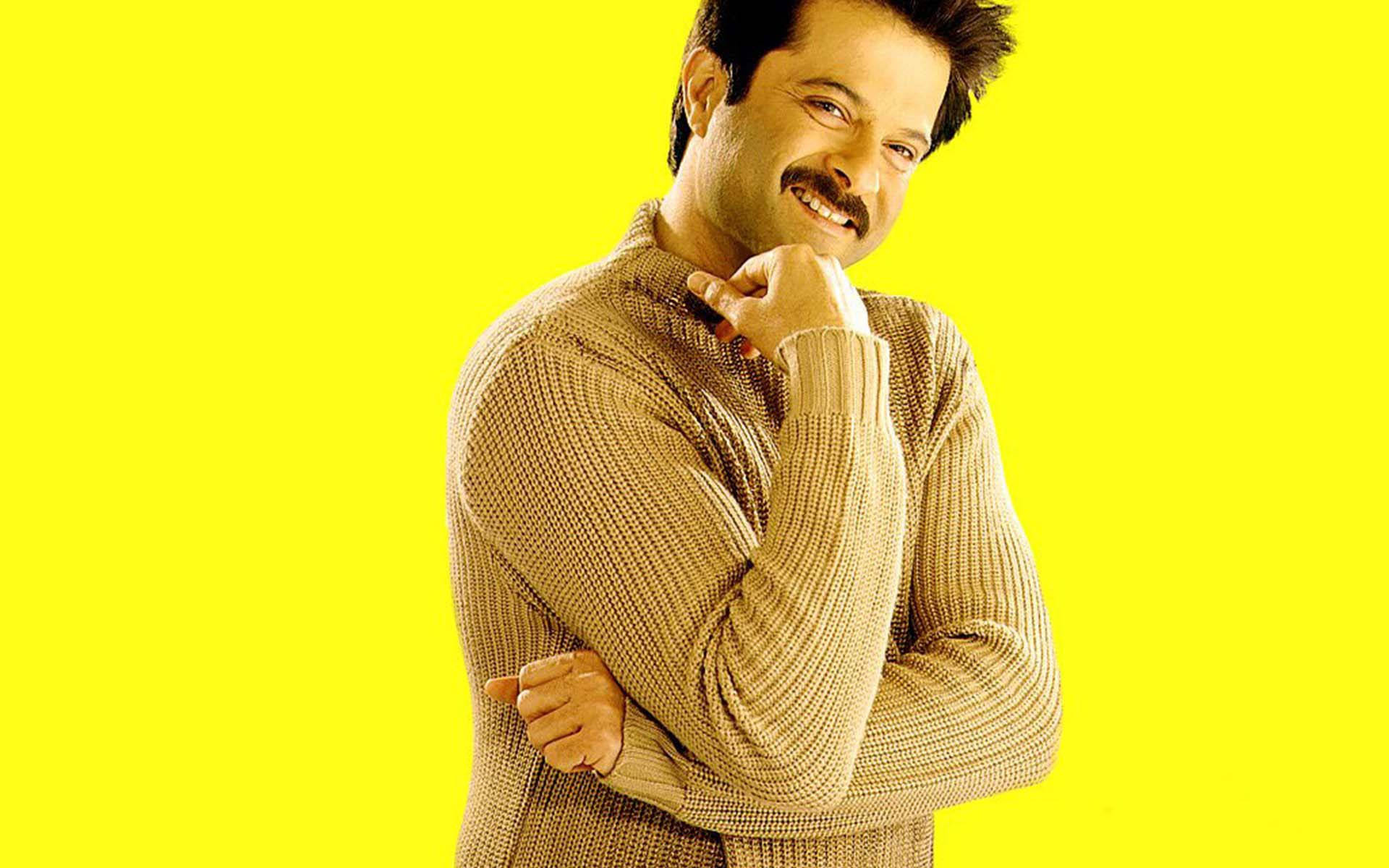 anil kapoor 桌布 壁紙 — 一名身穿米色毛衣的男子在明亮的黃色背景前，一手托腮，自信地微笑。