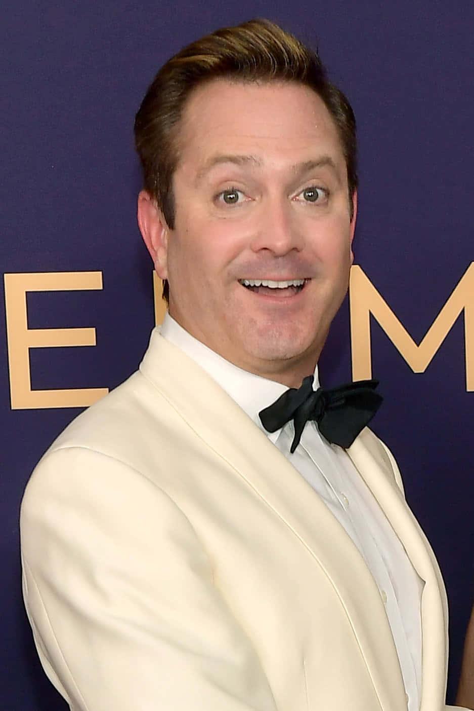 thomas lennon 桌布 壁紙 — 一名身穿米色燕尾服、繫著黑色領結的男子，在帶有金色字樣的紫色背景前燦爛微笑。