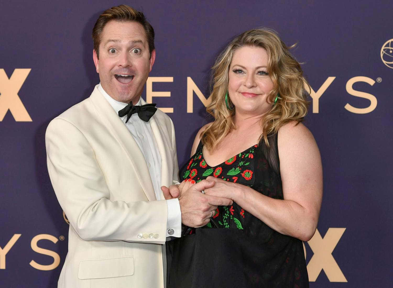 thomas lennon 桌布 壁紙 — 一名身穿米色燕尾服的男士和一名身穿花卉連衣裙的女士在艾美獎紅地毯上以誇張的表情擺姿勢。